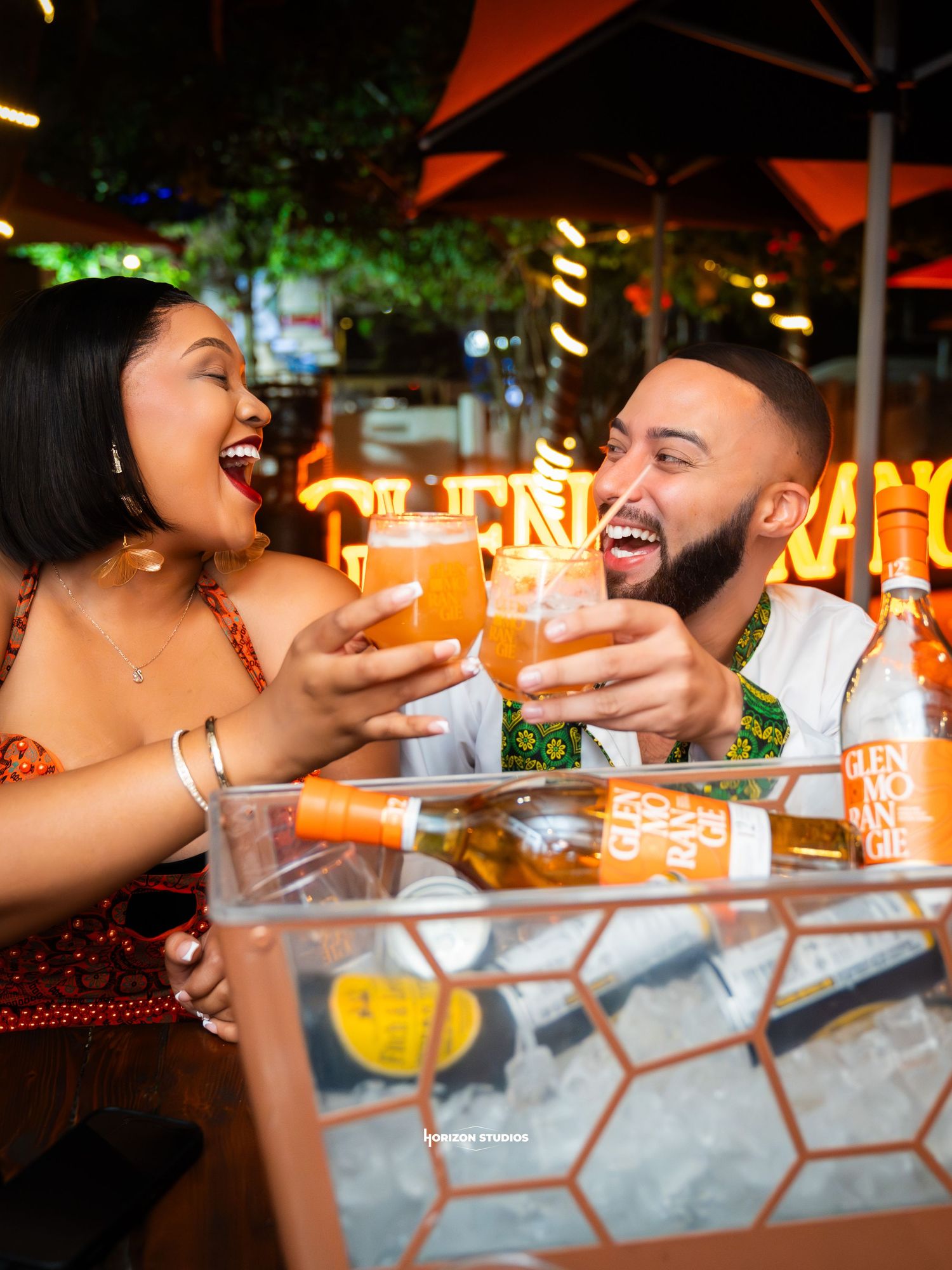 A joyful couple toasting with amber cocktails at an outdoor bar, surrounded by glowing neon lights at night.