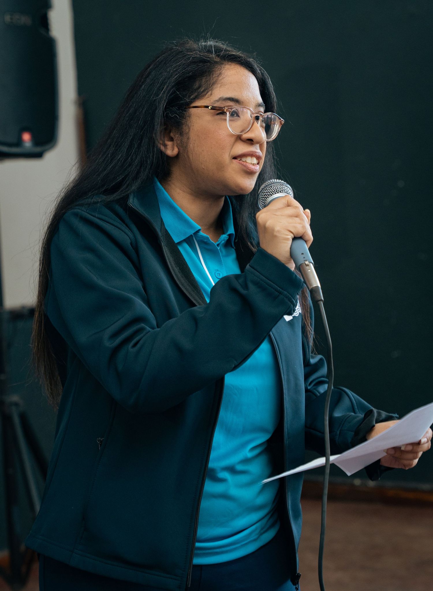 Young woman with glasses speaking into a microphone, holding papers, wearing a teal jacket at a public event.
