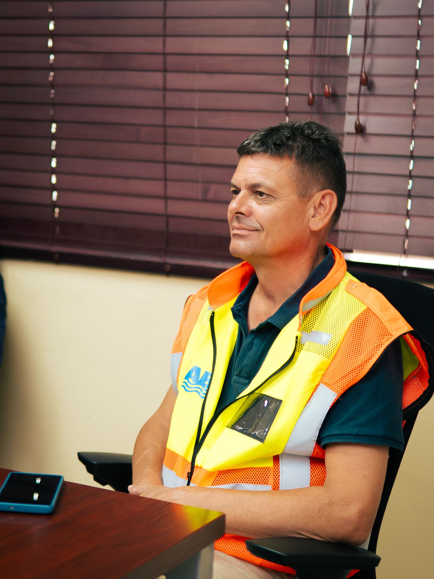 Man in high-visibility safety vest sitting at a desk in an office, looking attentive during a meeting.