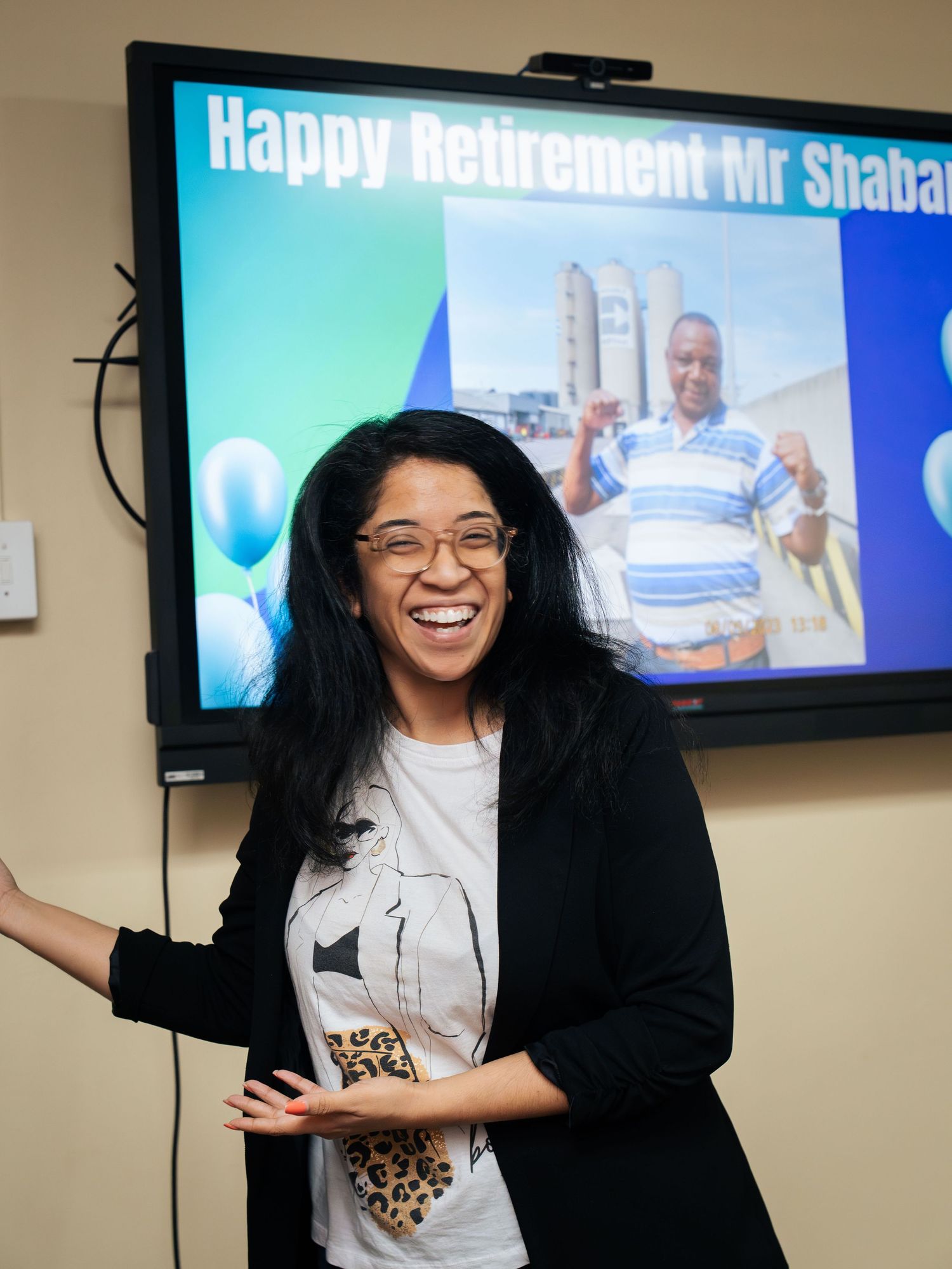 Woman smiling at a retirement party celebration with a Happy Retirement Mr Shaba slideshow displayed on screen behind her.