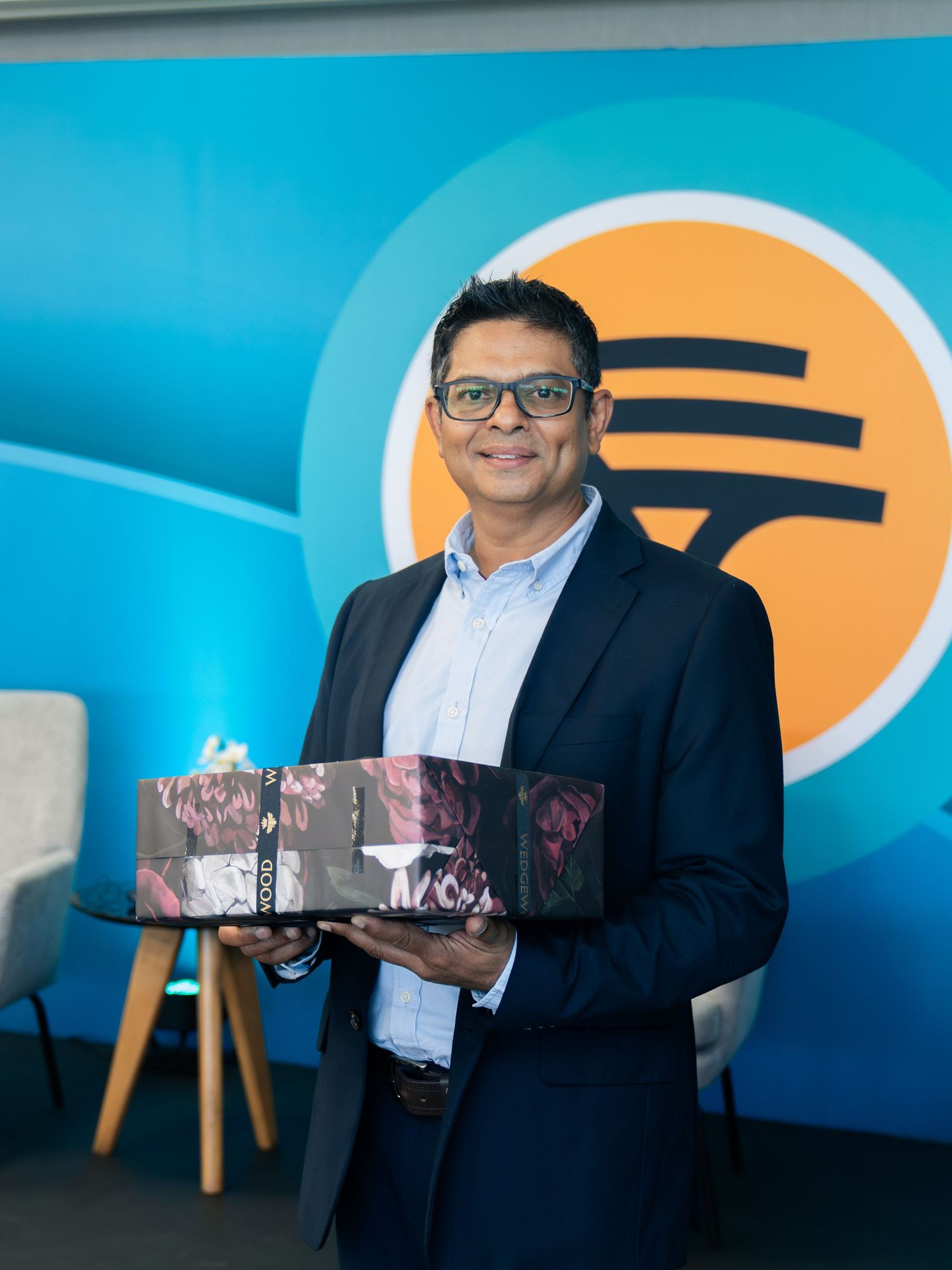 A smiling man in a navy blazer holds a decorative box in front of a blue and orange branded backdrop.