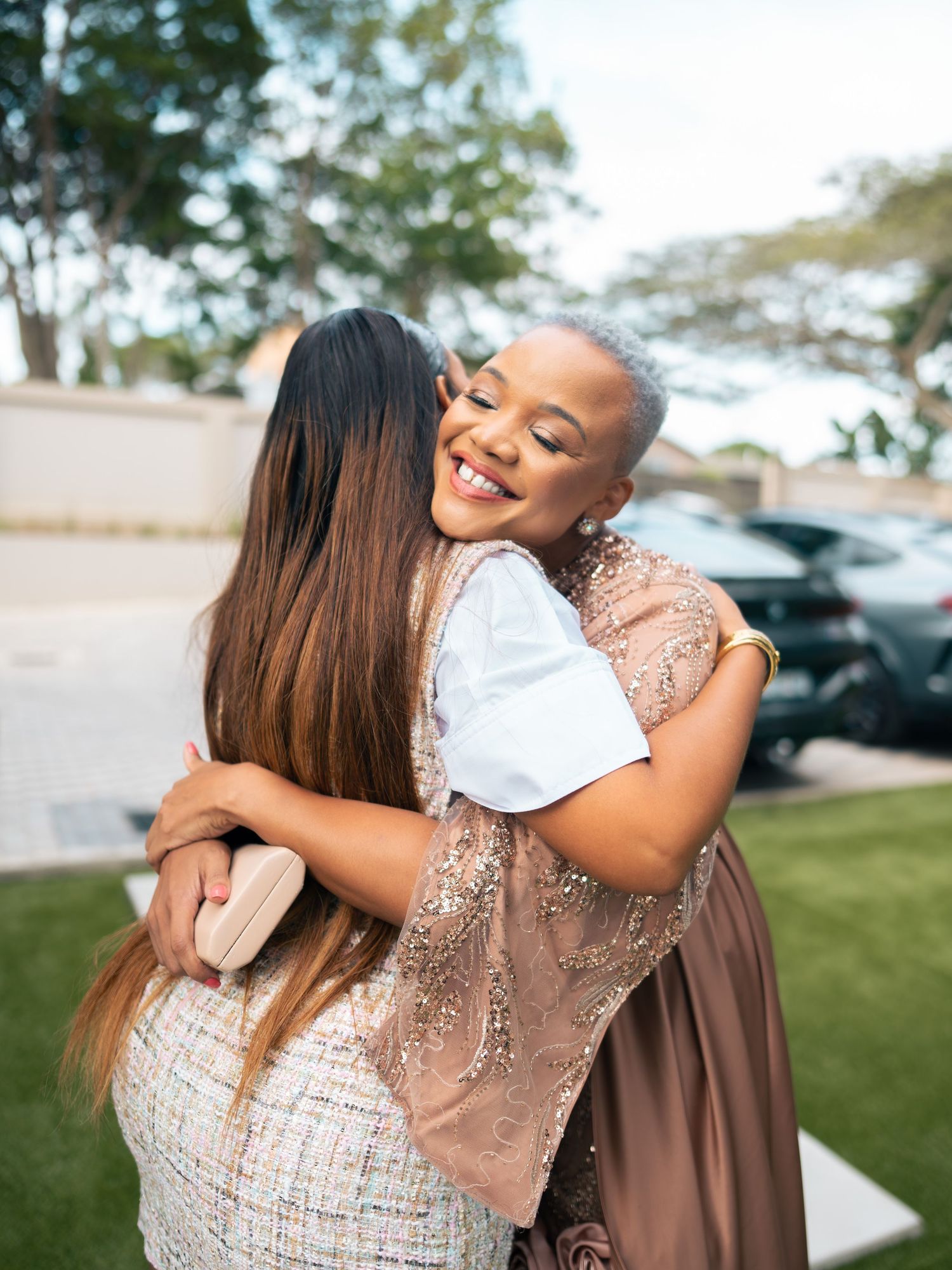 Two women sharing a warm embrace outdoors, smiling joyfully, dressed in elegant attire at a formal event.
