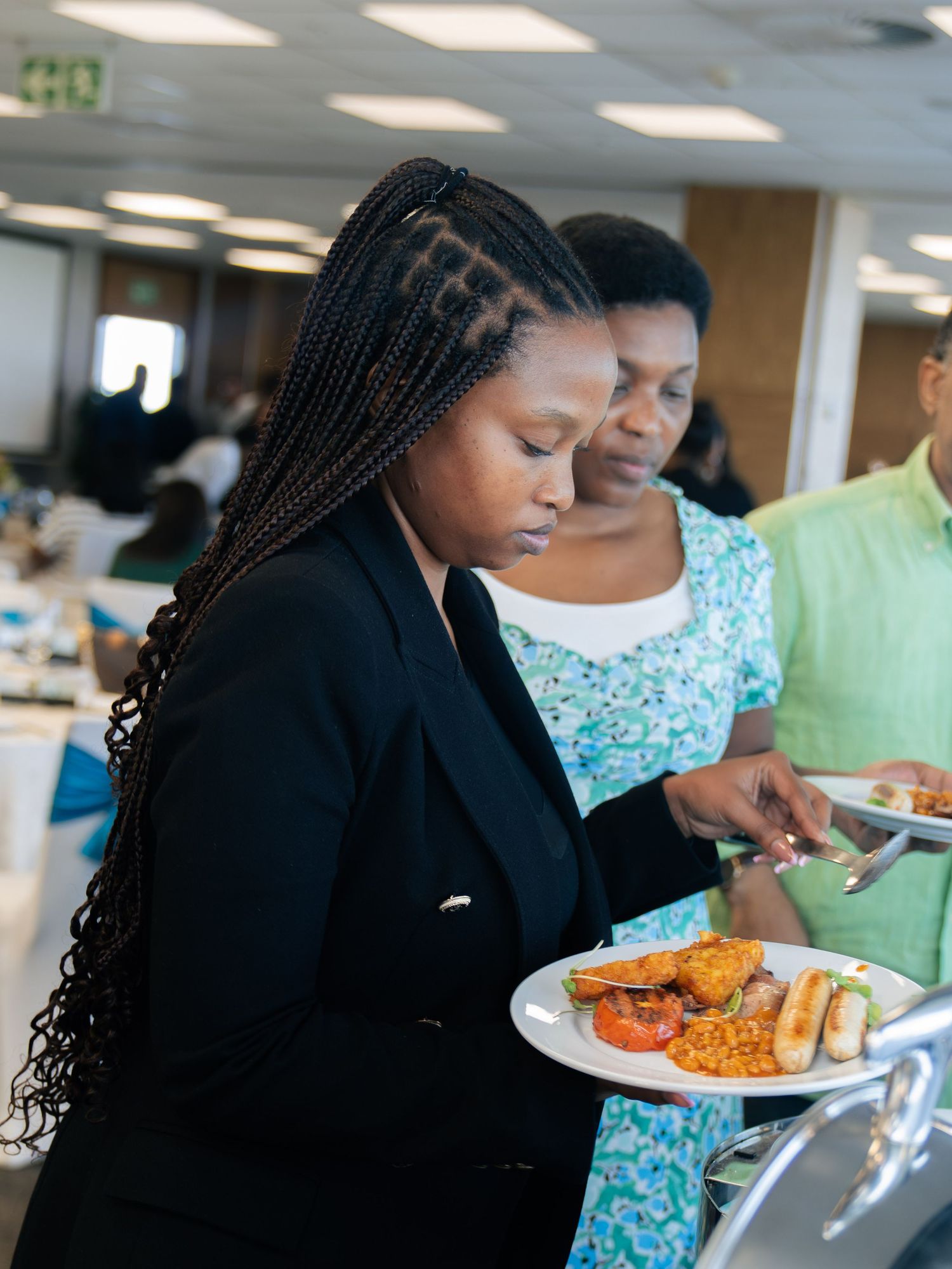 Two women serving themselves at a buffet, one in a black blazer with braided hair holding a plate of food.