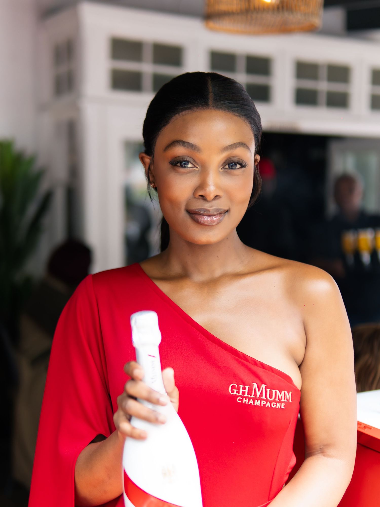 Woman in red one-shoulder G.H. Mumm Champagne dress holding a bottle at an outdoor event.
