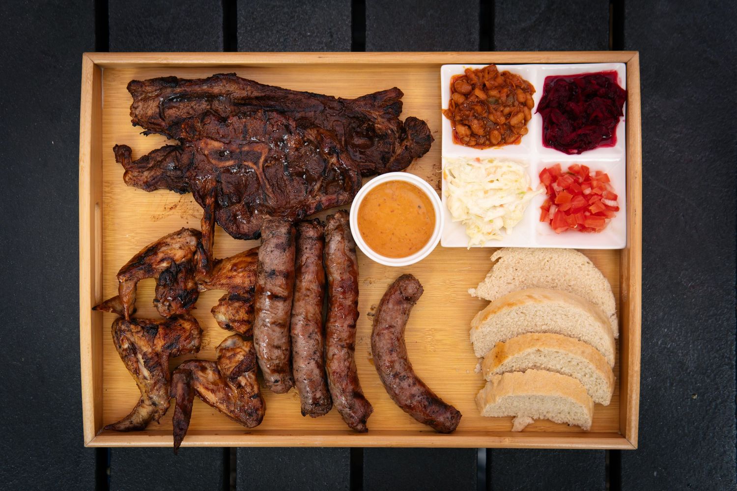 A wooden tray with BBQ meats, sausages, roasted mushrooms, bread, and condiments including sauce and fresh toppings.