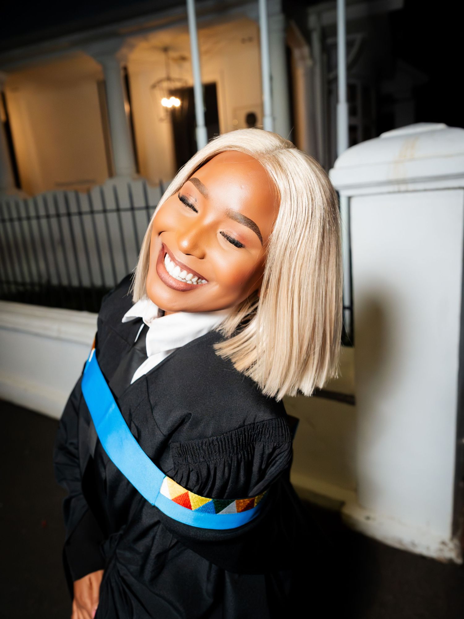 Smiling graduate in black gown with blue sash and blonde bob hairstyle posing outdoors at night.