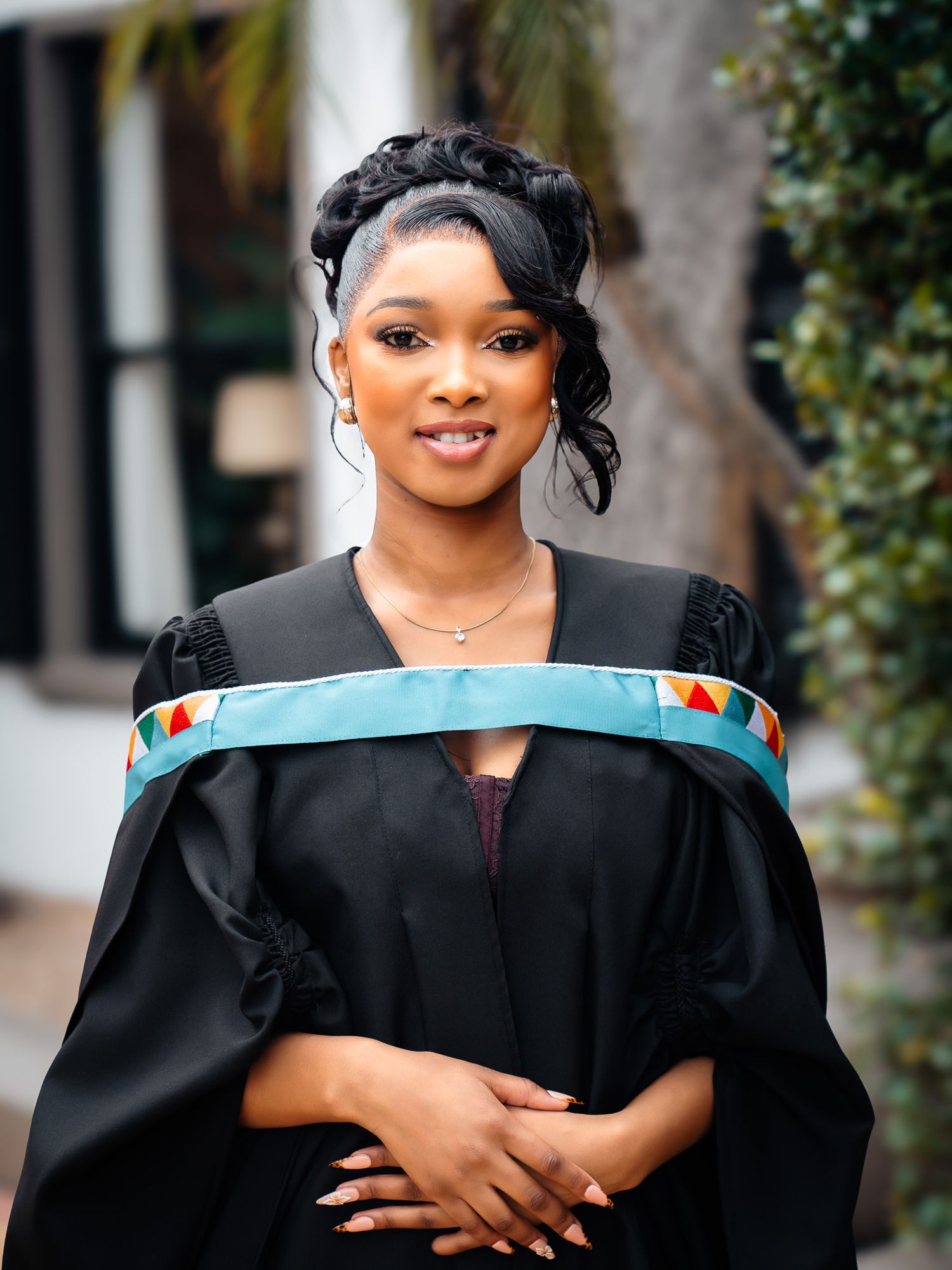 Young woman in black graduation gown with colorful hood smiles proudly outdoors on her graduation day.