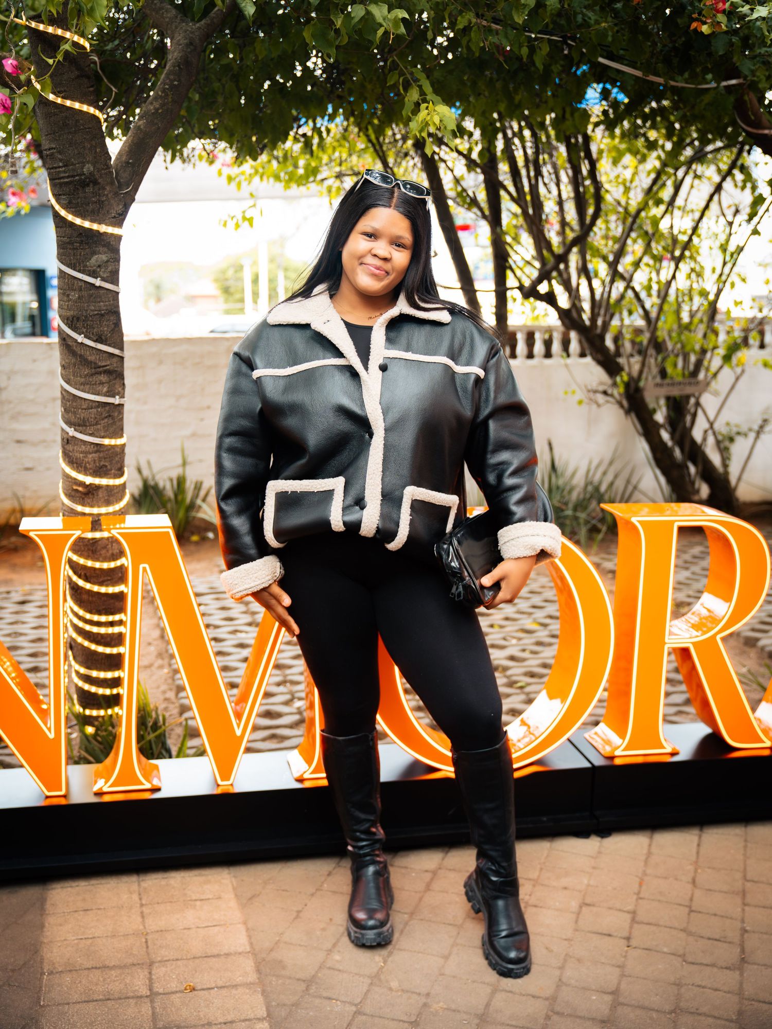Woman in black leather jacket and leggings poses confidently in front of illuminated orange letters outdoors.
