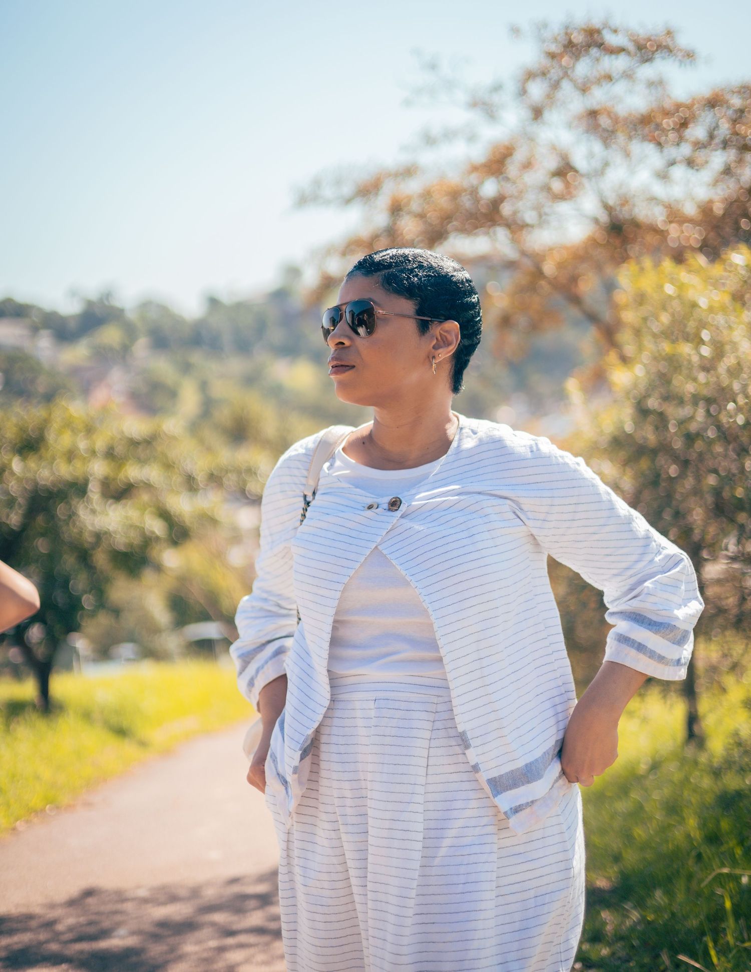 Woman in striped outfit and sunglasses standing on a sunny park path surrounded by lush green trees.