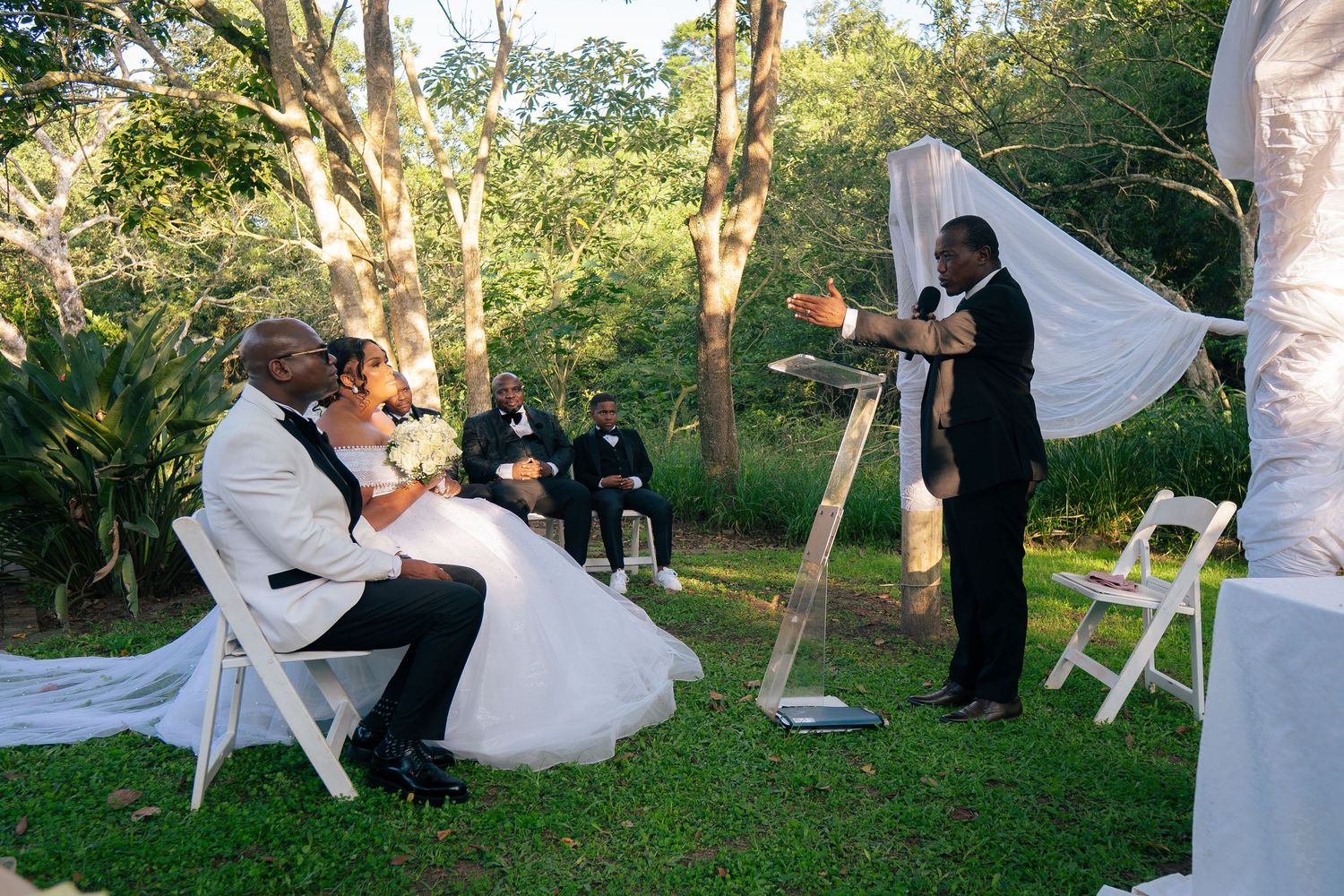 Outdoor wedding ceremony with couple seated before officiant, white chairs, draped arch, and lush green trees backdrop.