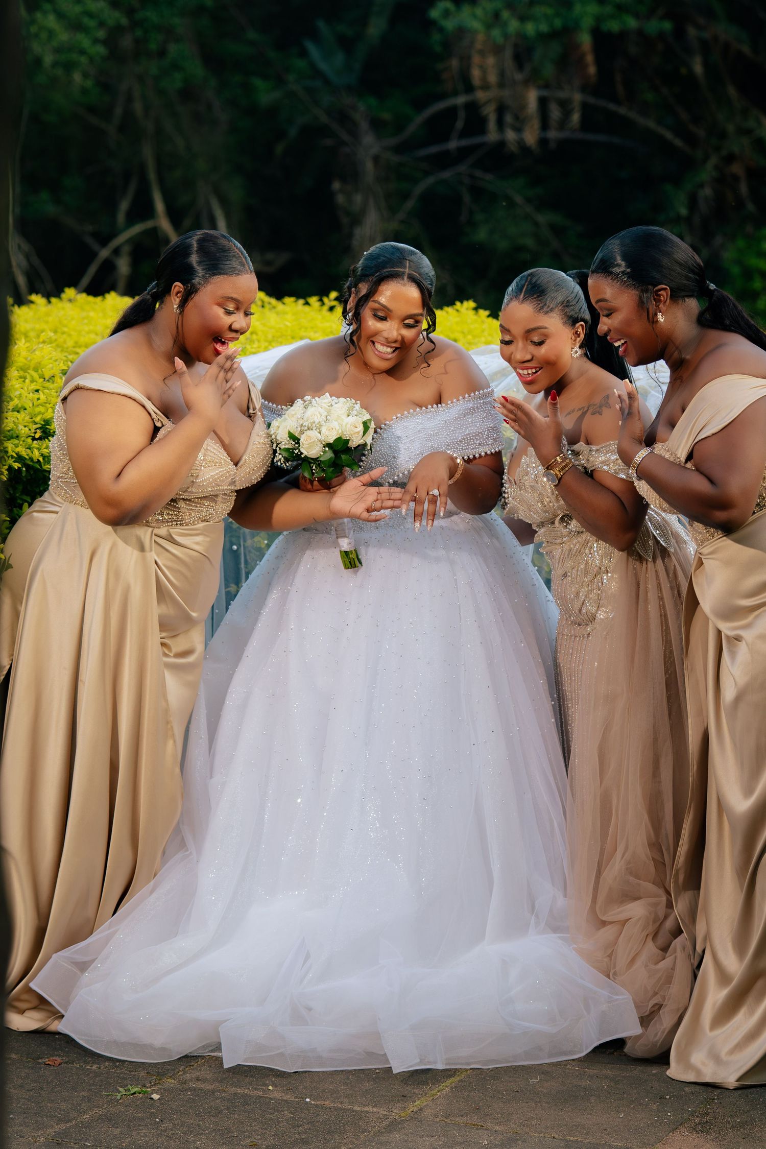 Bride in white ball gown holds bouquet surrounded by four bridesmaids in gold dresses, all smiling outdoors.