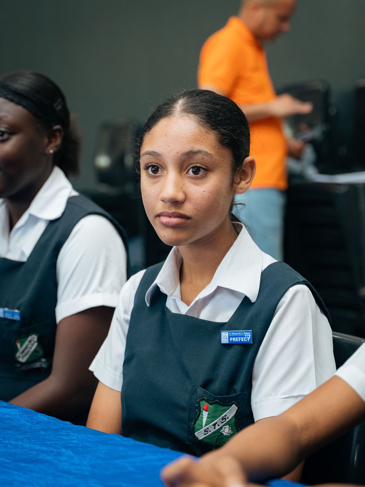 Young student in green and white school uniform sits at a blue table in a classroom setting.
