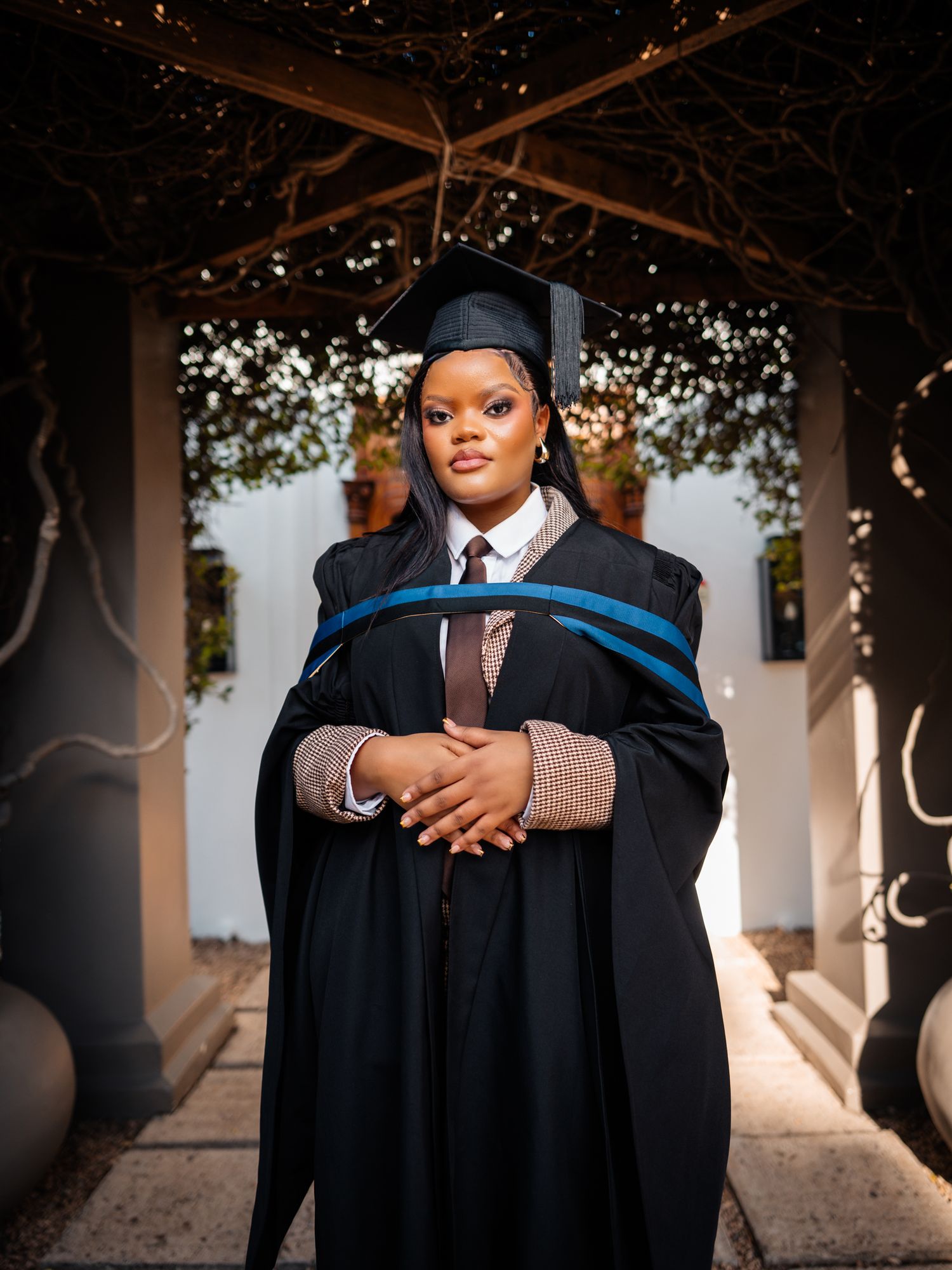 A proud graduate in black cap and gown with blue sash poses outdoors under decorative branches.