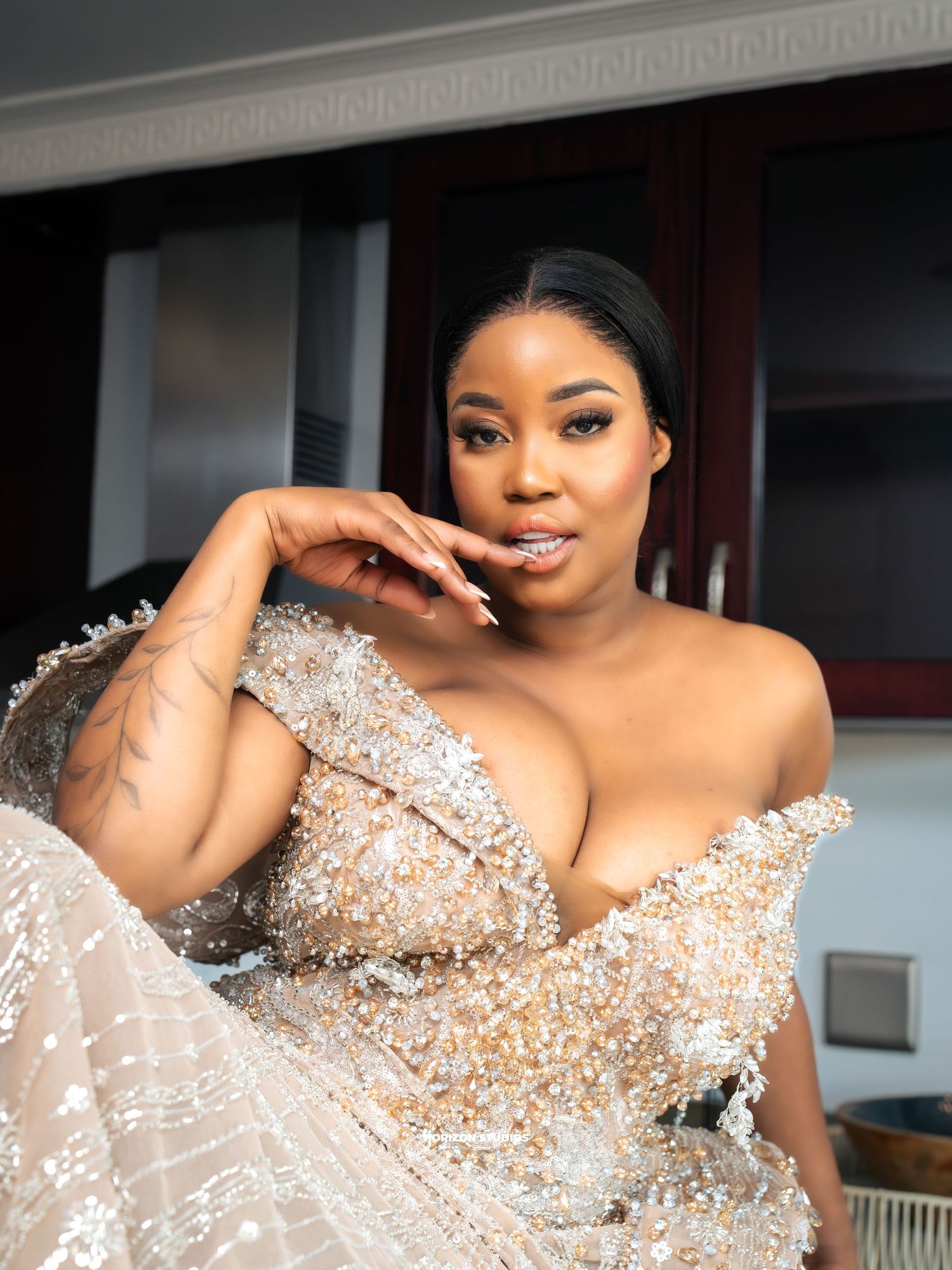 Woman in elegant beaded off-shoulder gown posing confidently indoors, showcasing glamorous formal attire.