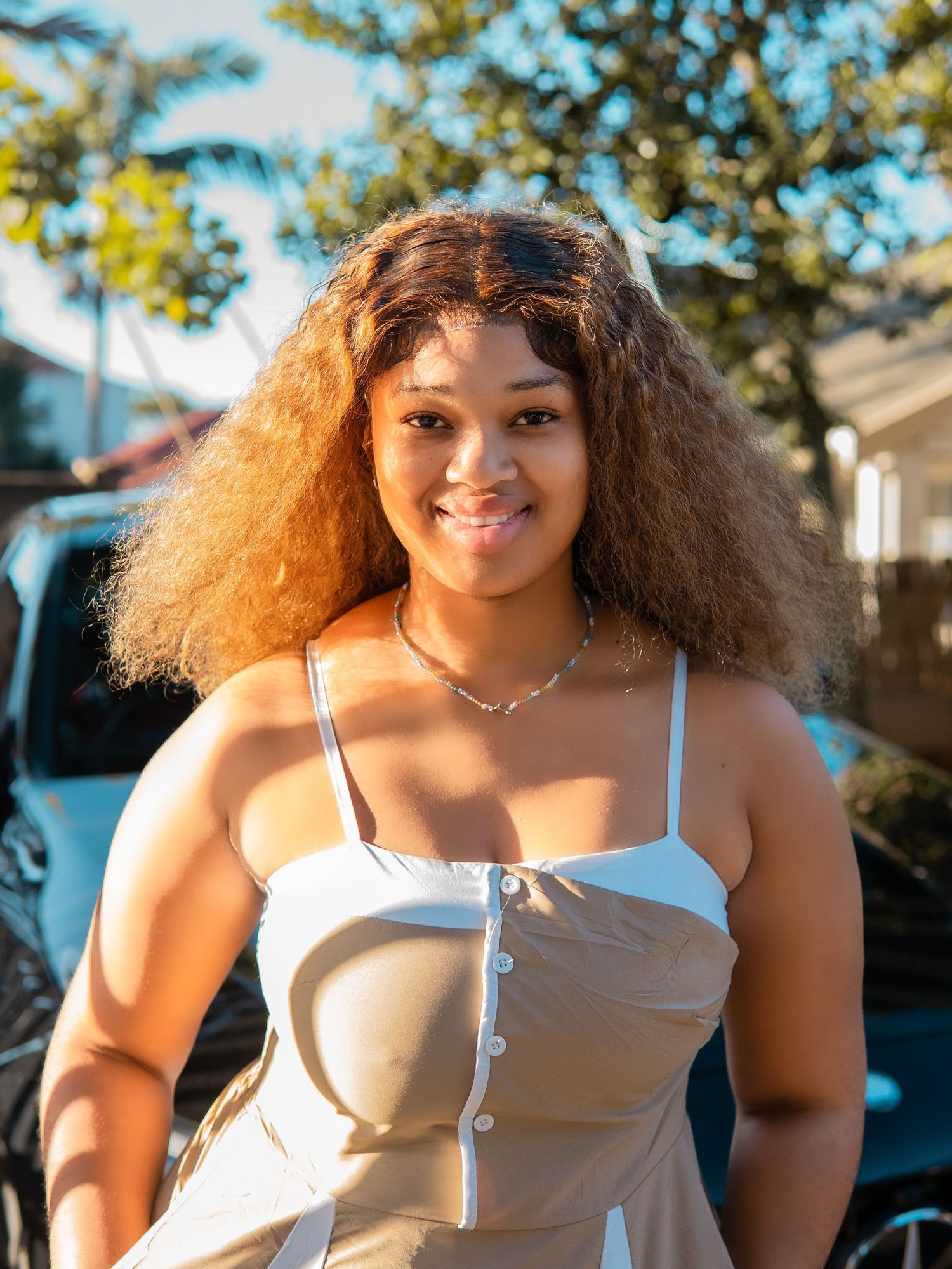 Young woman with curly hair smiling outdoors in a white button-up dress, with trees and a vehicle in the background.