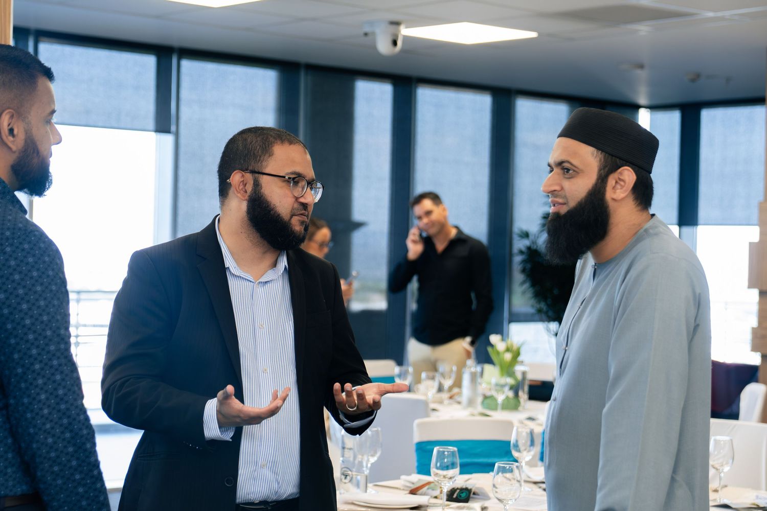 Men in formal attire engaged in conversation at an indoor dining event with tables set in the background.