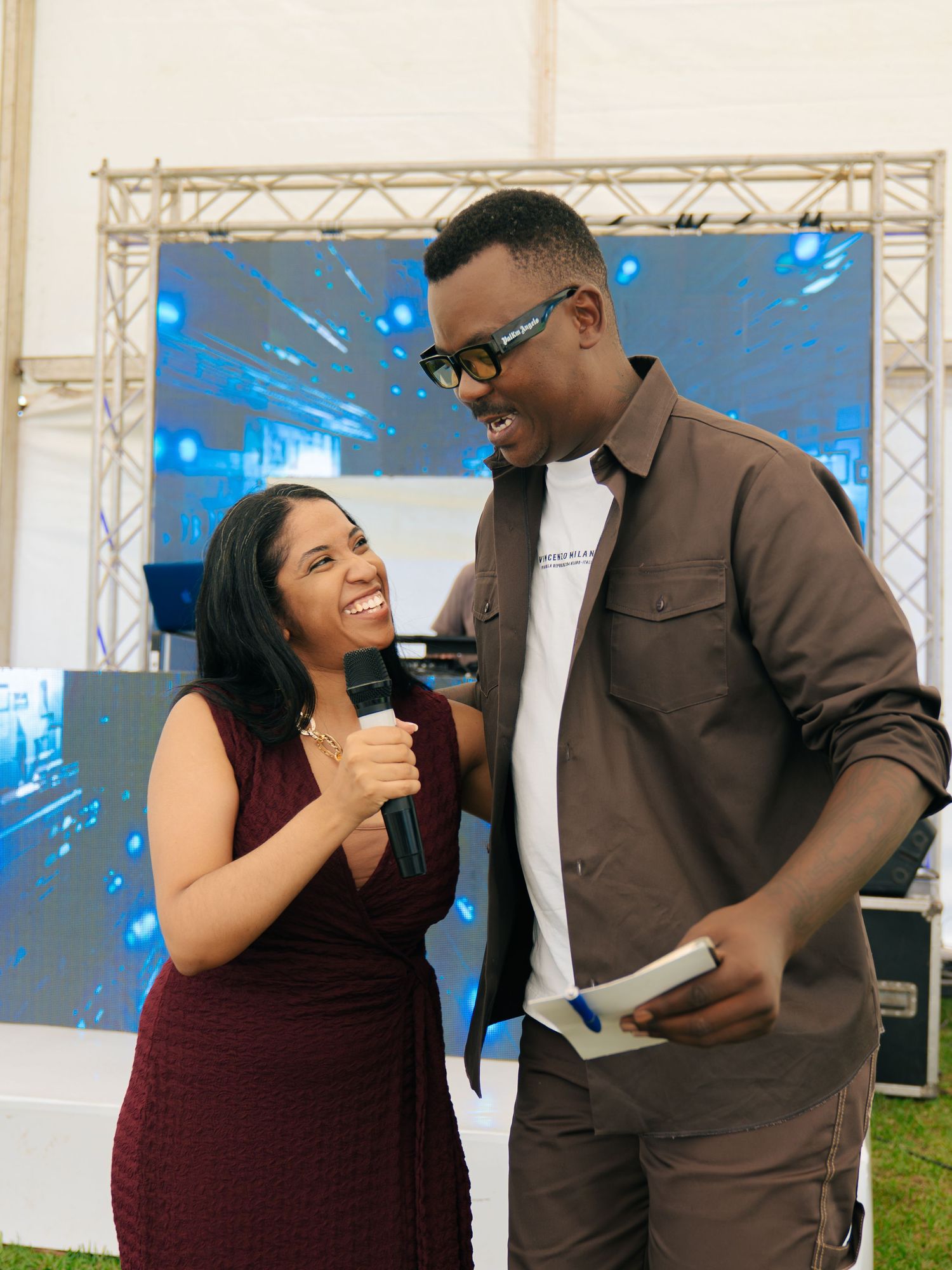 A woman in a burgundy dress holds a microphone while interviewing a man in a brown shirt at an outdoor event.