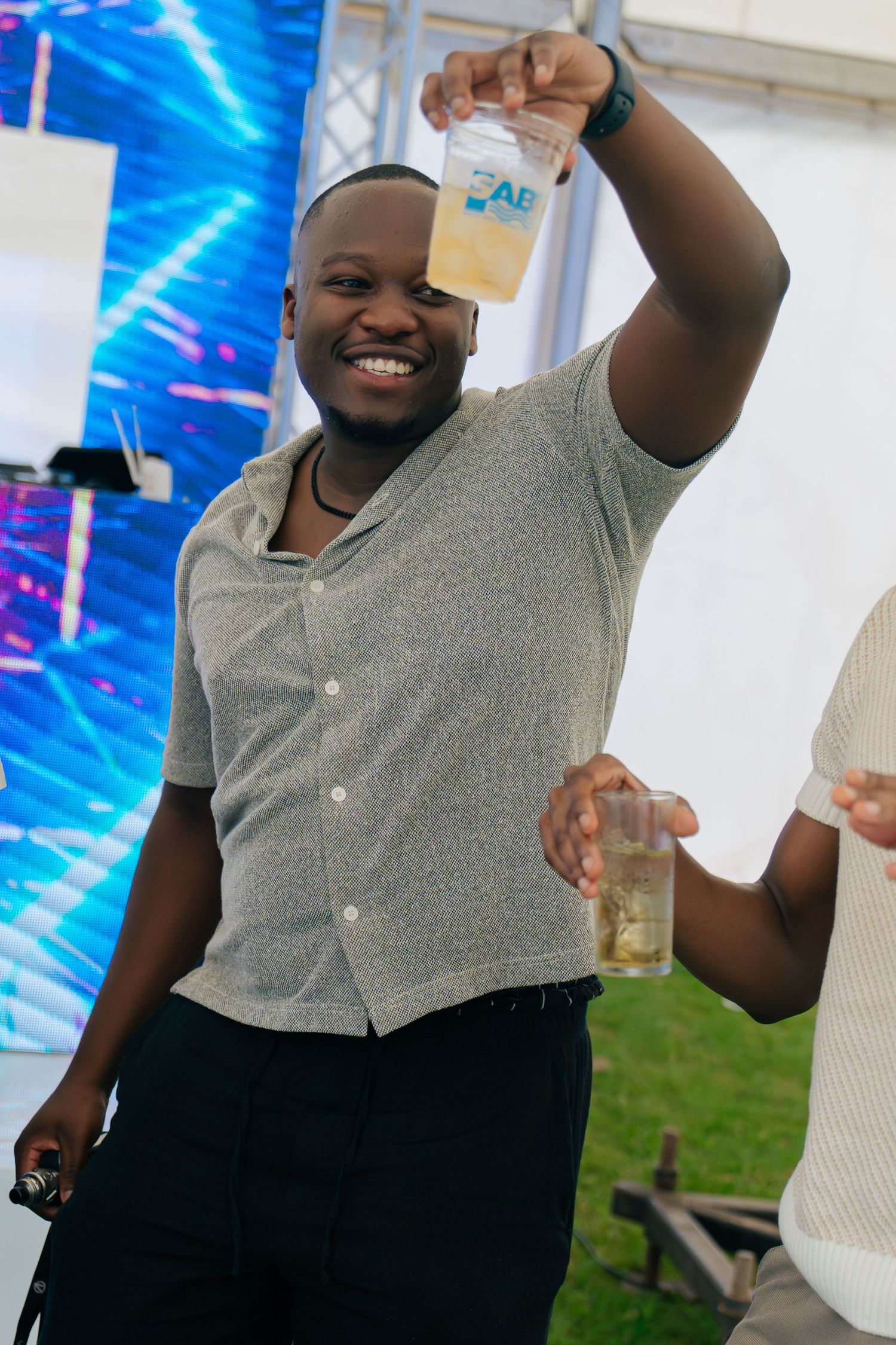 Smiling man holding up a plastic cup of beer at an outdoor festival event.