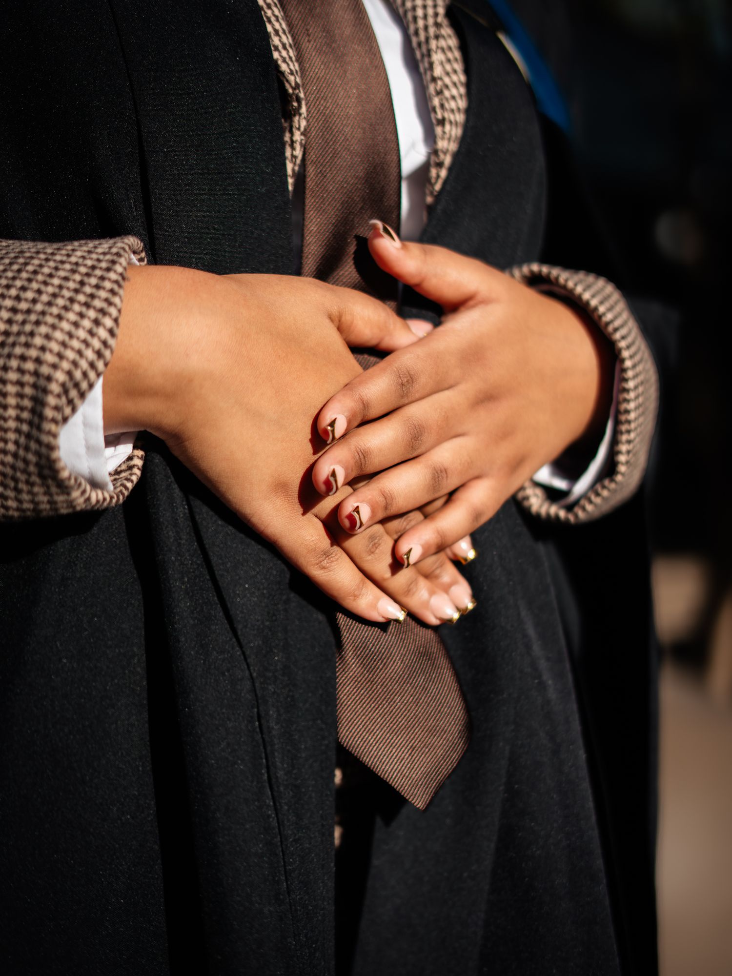 Close-up of hands with dark red nails, wearing a black coat and brown tie, clasped together in natural light.