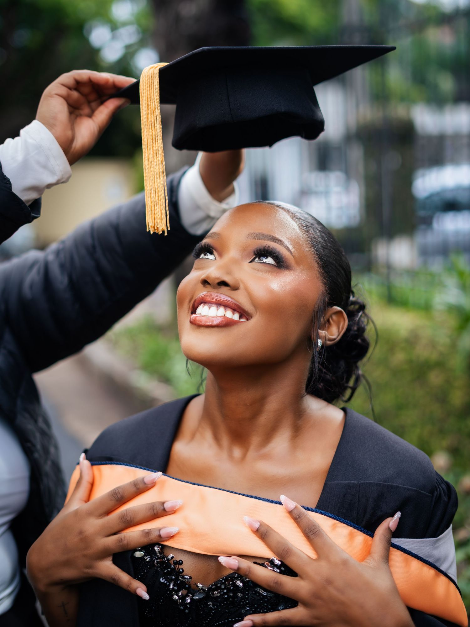 Smiling young Black woman in graduation cap and gown receiving her mortarboard outdoors.
