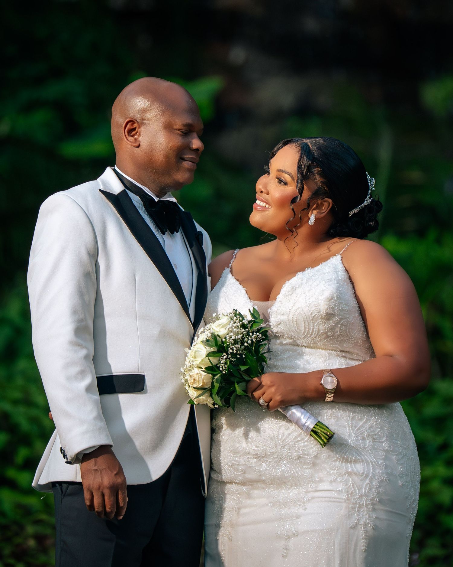 A couple in wedding attire smiles at each other, bride holding white bouquet, groom in white tuxedo, outdoors.