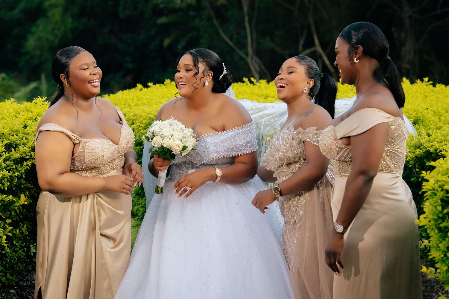 Bride in white gown with bridesmaids in champagne dresses laughing outdoors amid lush green hedges.
