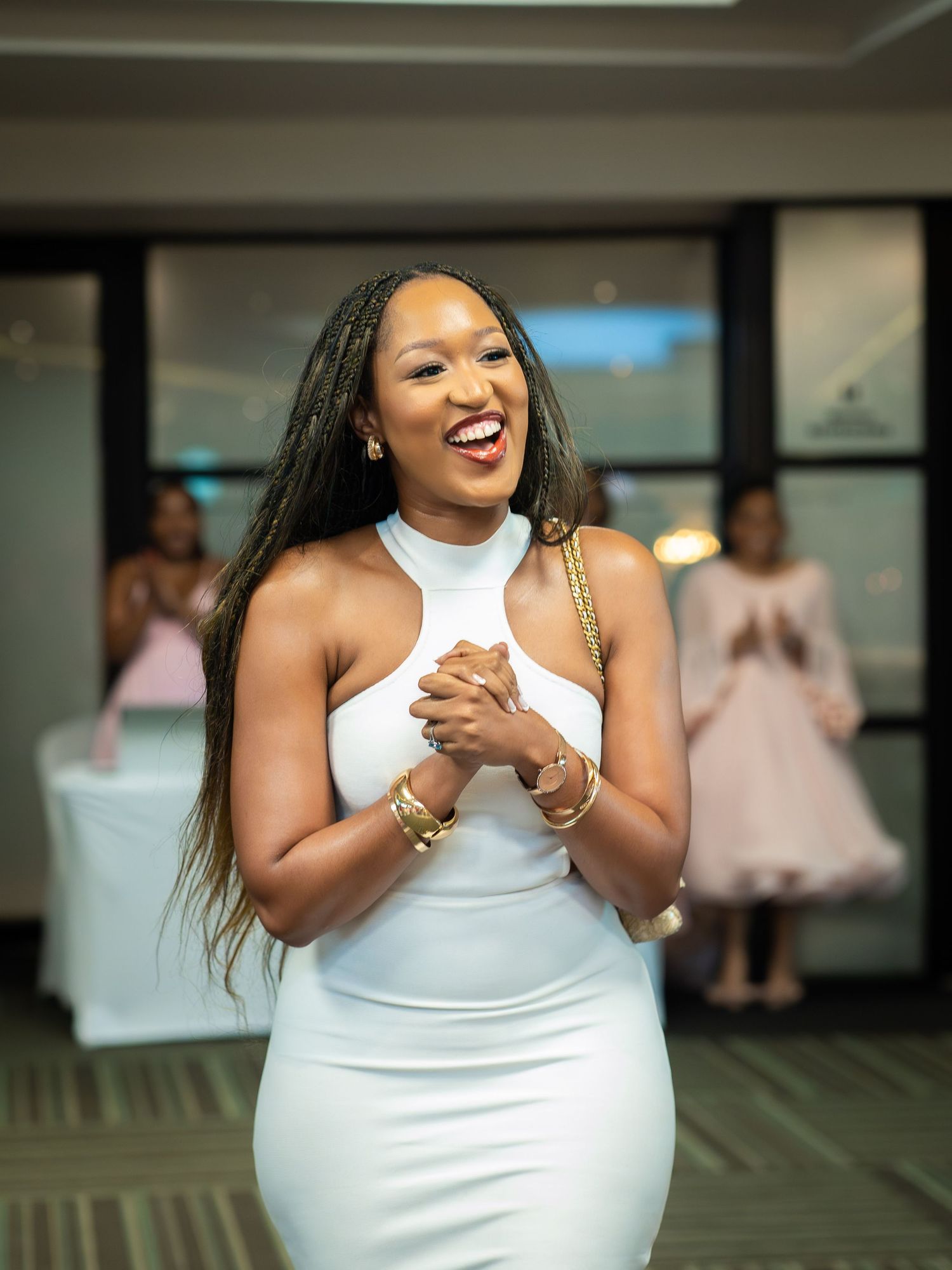 A joyful woman in a white halter dress laughing at an elegant event, wearing gold jewelry and braided hair.