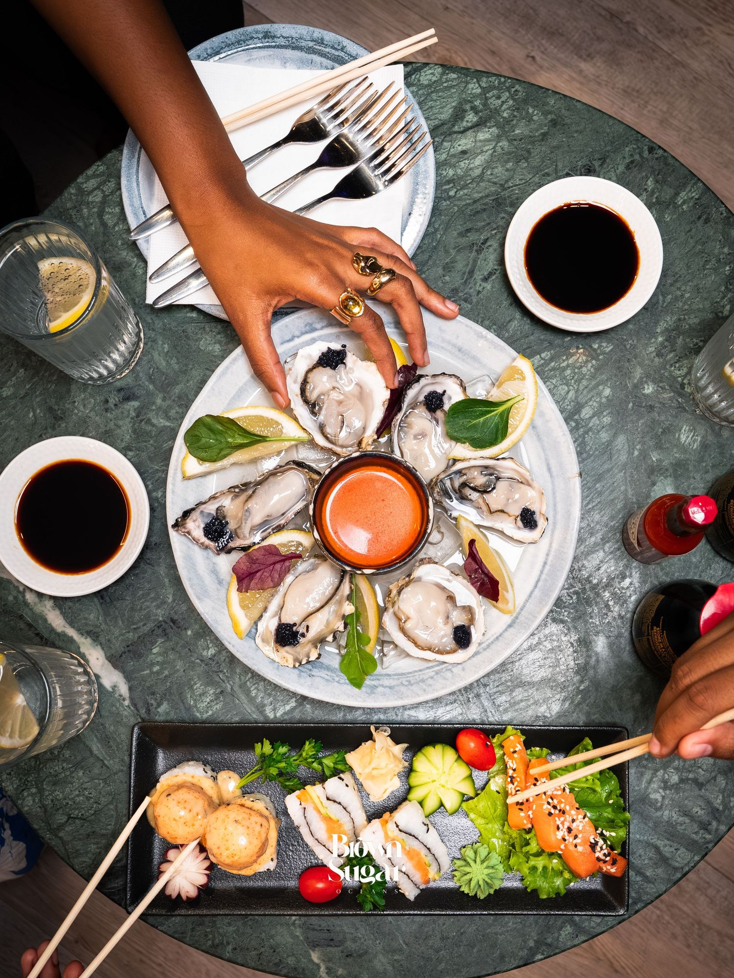 Overhead view of oysters on ice with dipping sauce and sushi platter on a marble table.