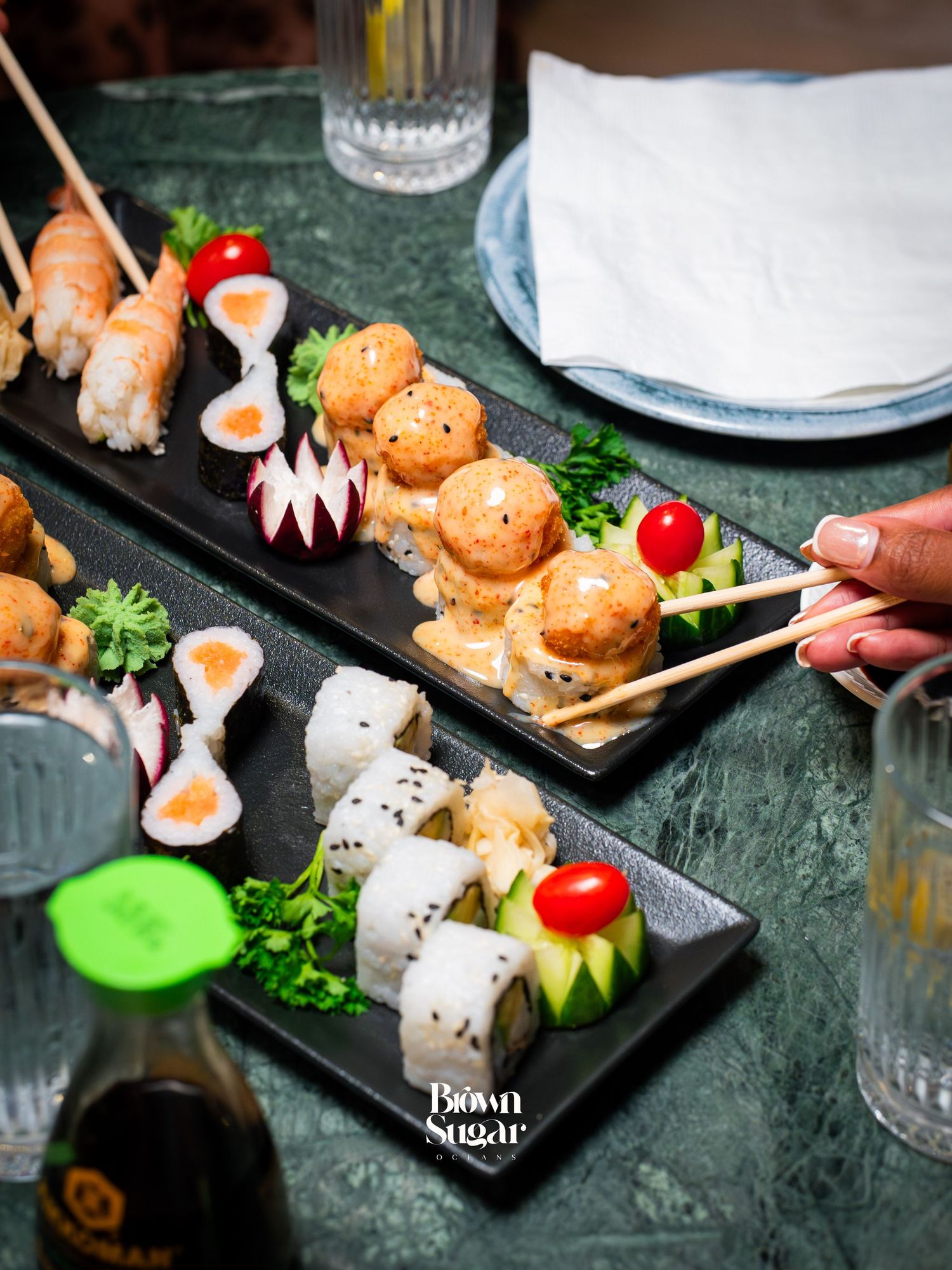 Two black platters of sushi rolls and salmon skewers garnished with cherry tomatoes and greens at Brown Sugar restaurant.