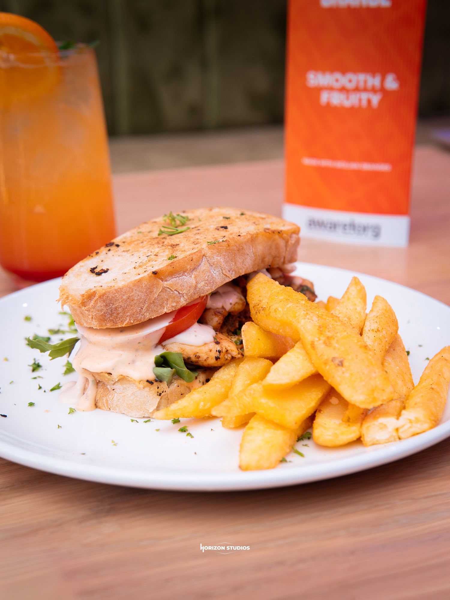 A sandwich with chicken, tomato, and sauce served with golden fries on a white plate beside a fruity orange drink.
