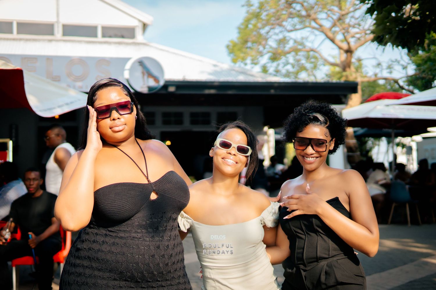 Three stylish women wearing sunglasses pose together outdoors at a social event on a sunny day.