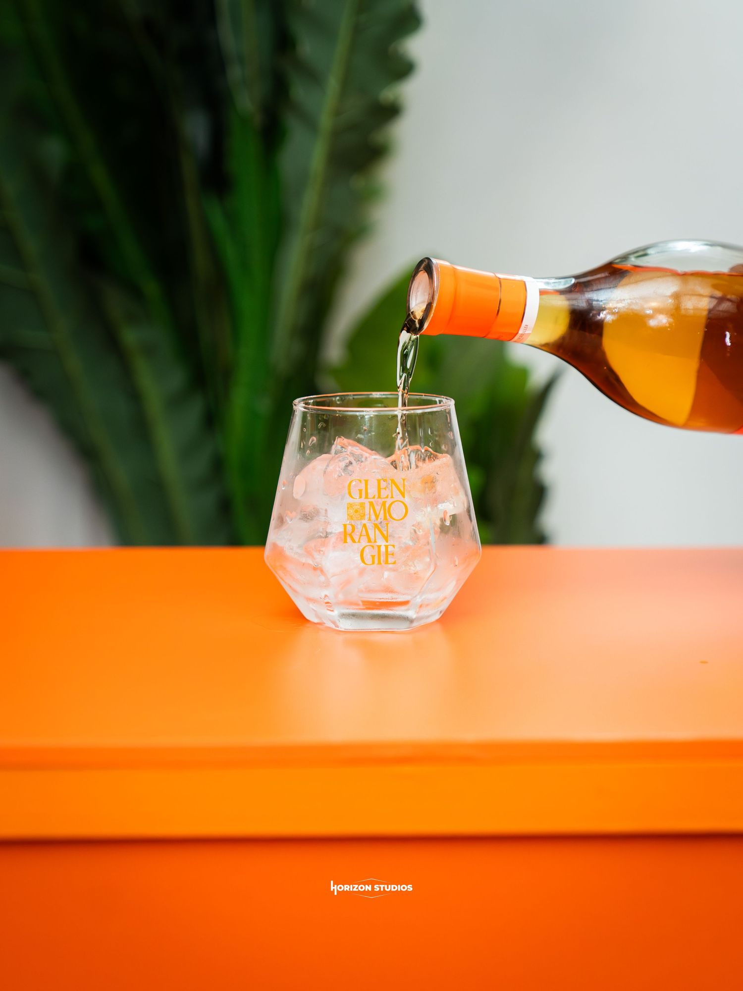 Glenmorangie whisky being poured into a branded stemless glass on an orange surface with green plants.