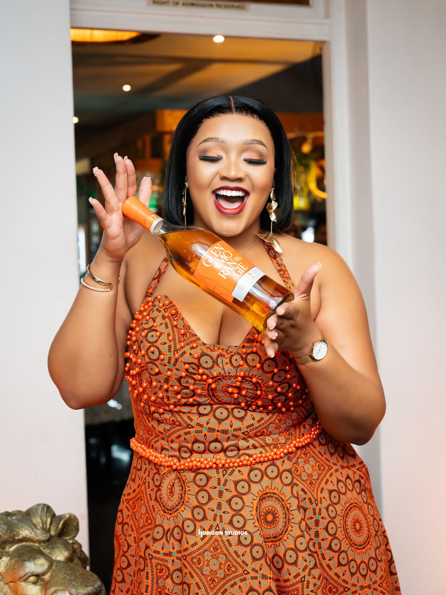Woman in orange beaded dress laughing while holding a bottle of liquor at an elegant event.