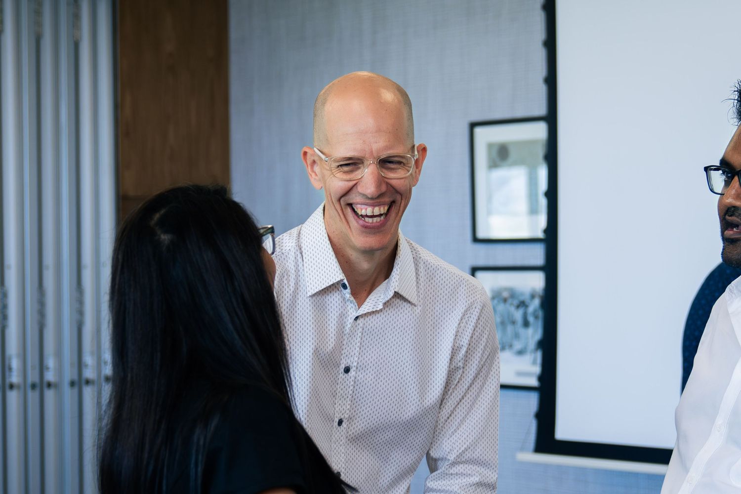 Bald man in white shirt laughing joyfully while networking with colleagues in a modern office setting.