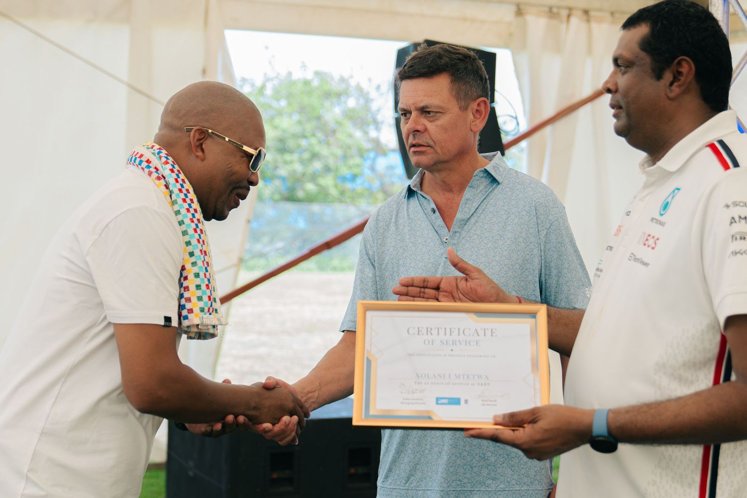 Three men shaking hands during a certificate presentation ceremony at an outdoor event under a white tent.