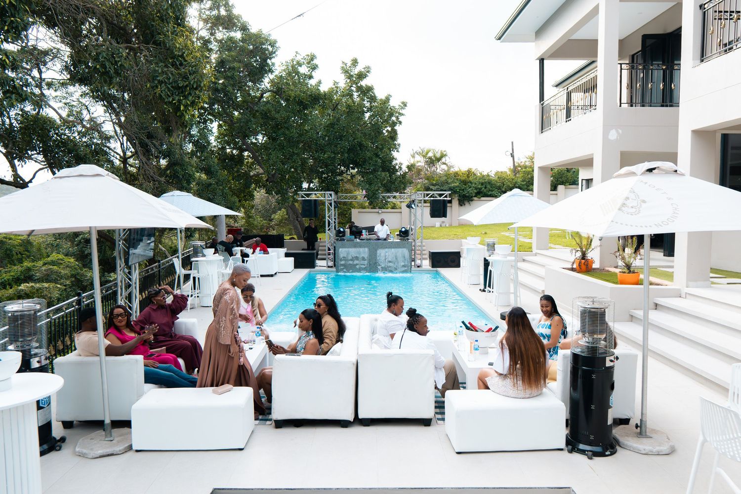 Elegant outdoor pool party with white lounge furniture, umbrellas, and guests socializing beside a luxury villa.
