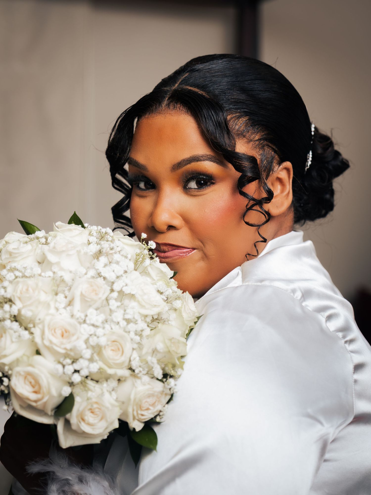 Bride holding white rose bouquet with elegant updo hairstyle and curls, wearing white satin robe on wedding day.