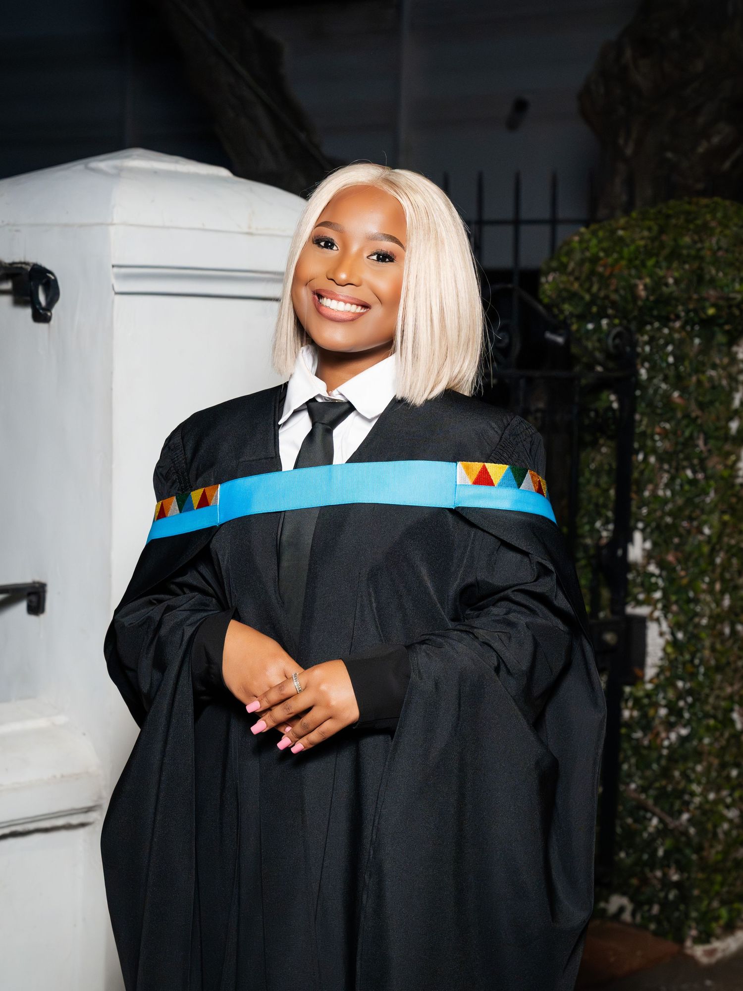 Smiling graduate in black academic gown with colorful sash poses proudly outdoors on graduation day.