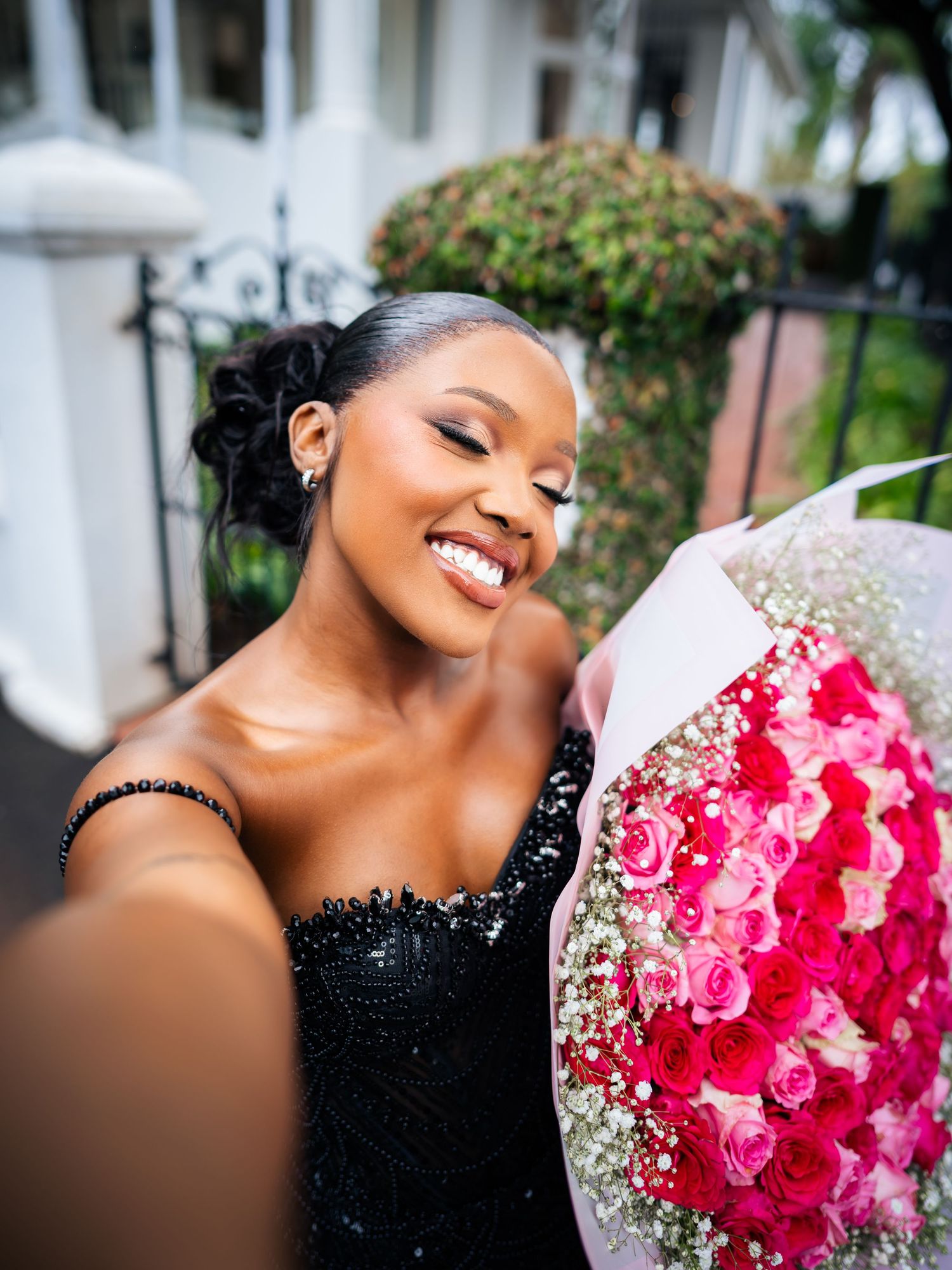 A smiling woman in a black sequin dress takes a selfie while holding a large bouquet of pink and red roses.
