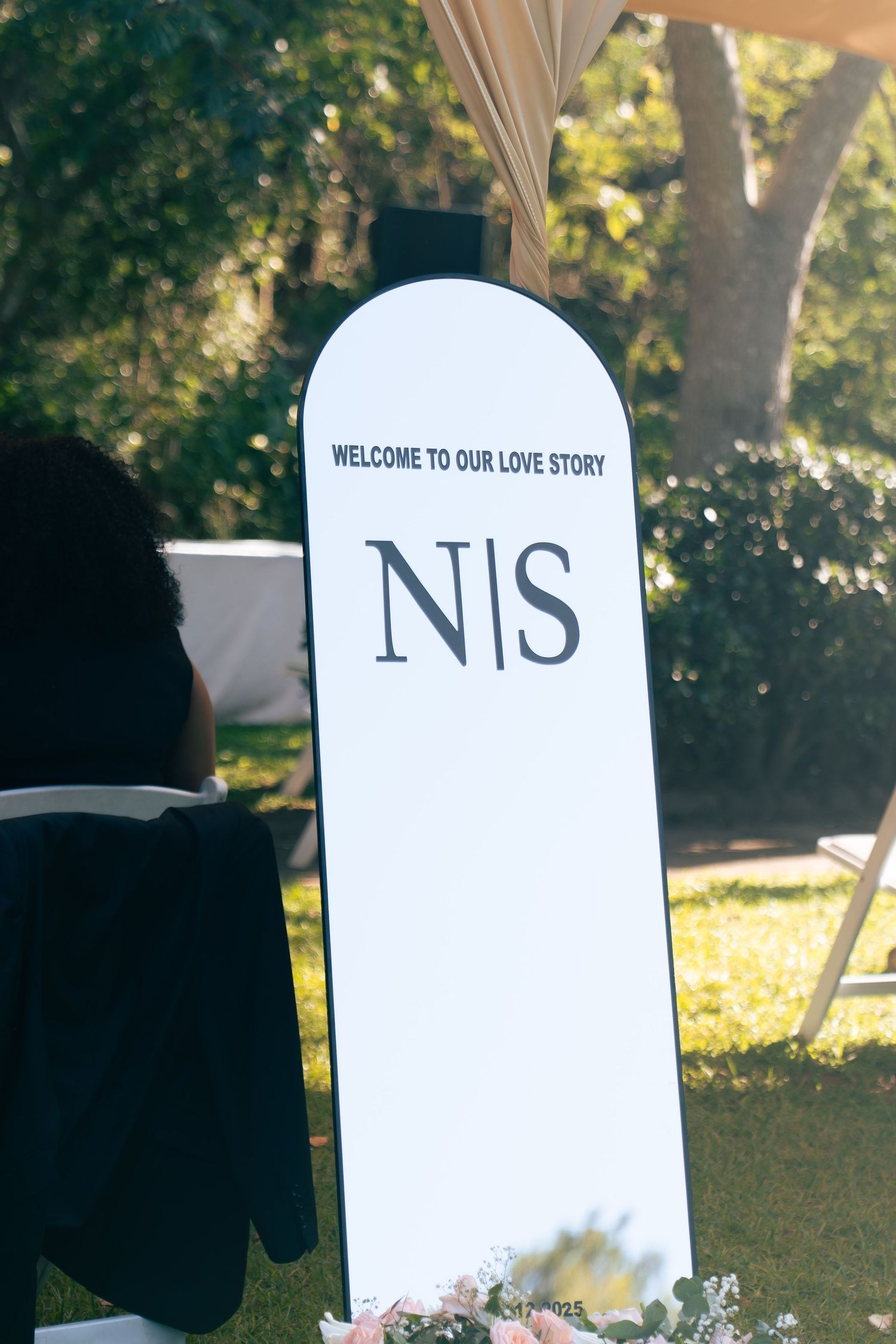 Outdoor wedding welcome sign reading Welcome To Our Love Story with initials N and S on white arch board.