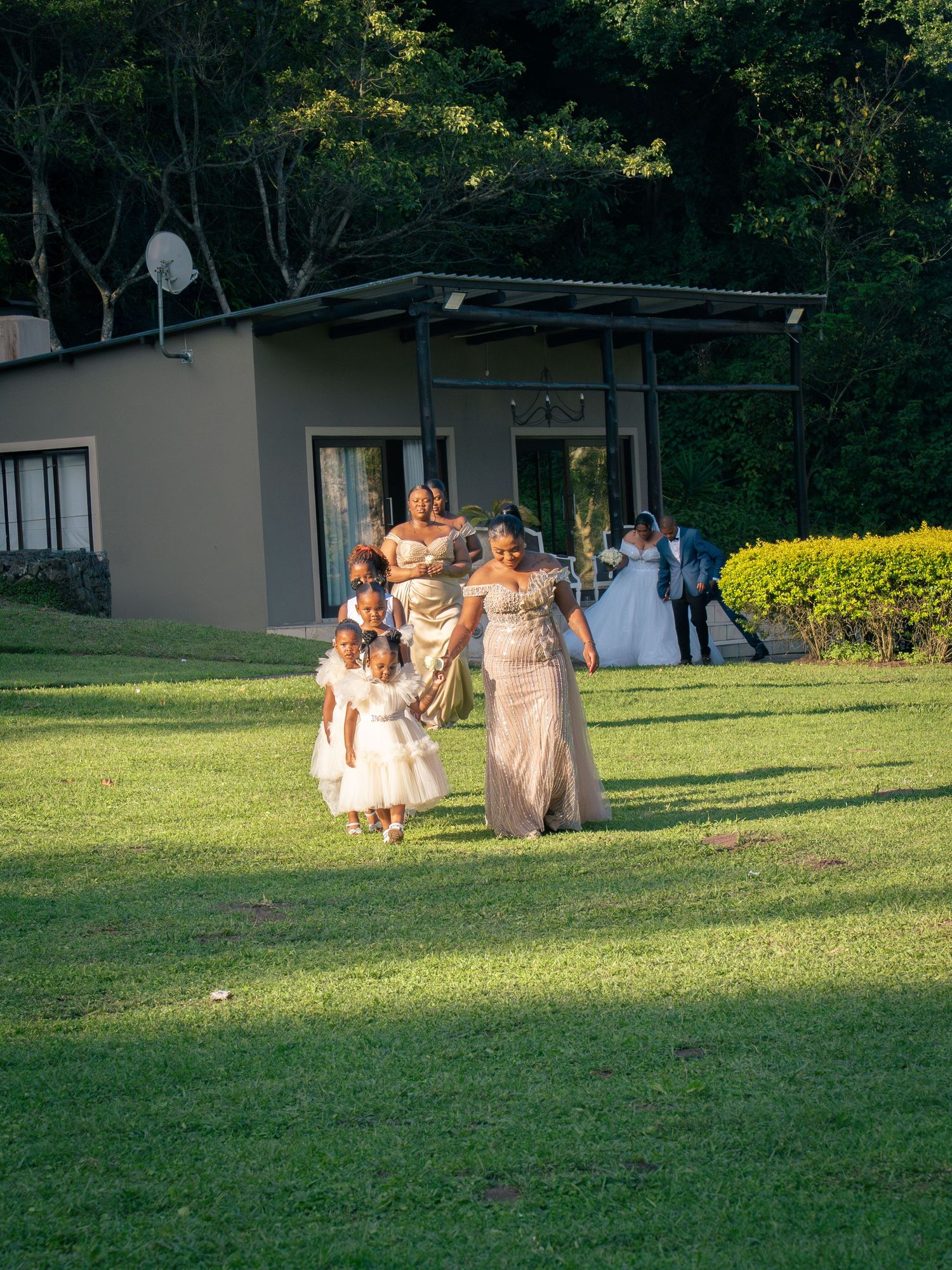 Wedding party walking across lush green lawn with flower girls and bridesmaids in elegant gowns near a modern venue.