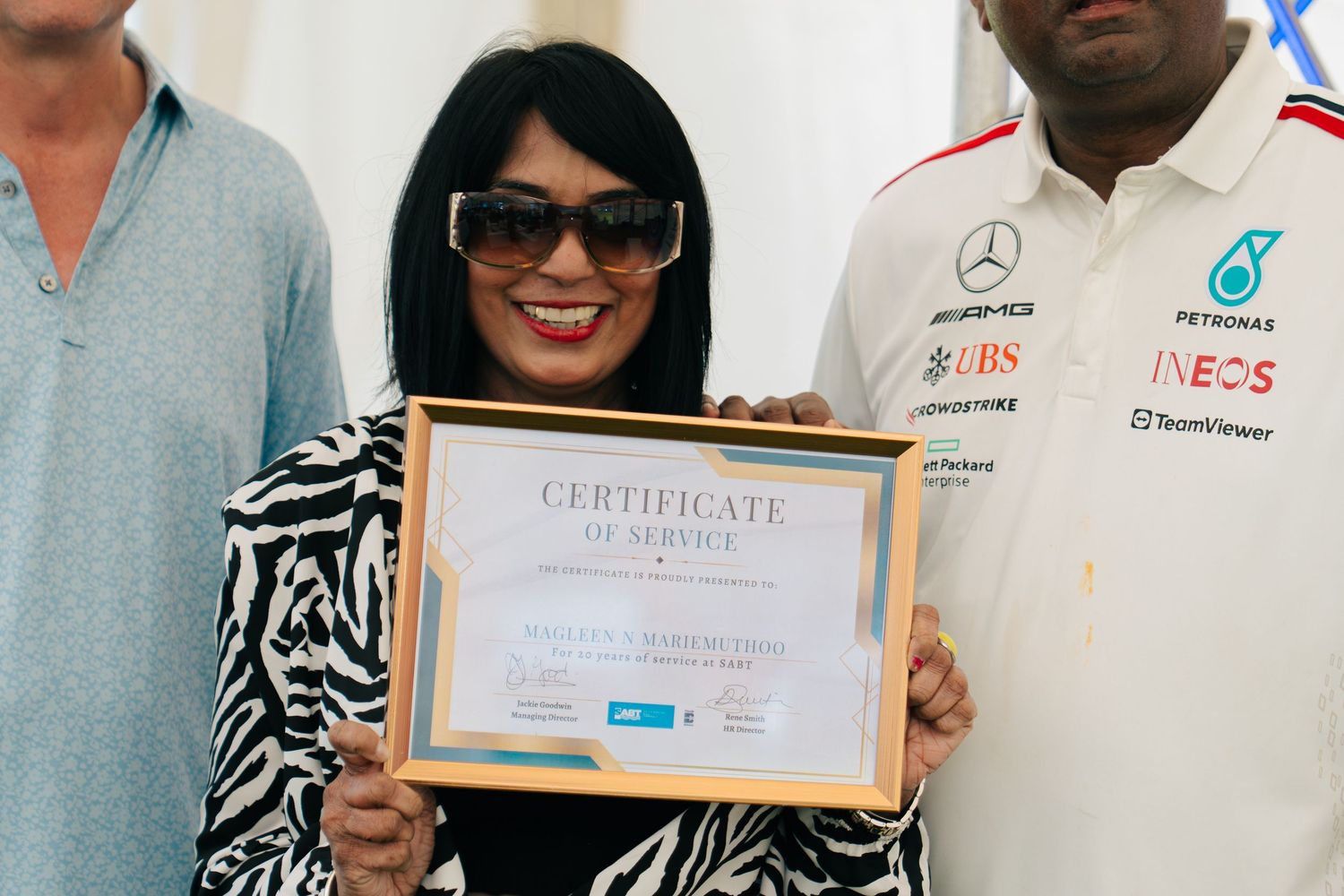 Woman proudly holding a Certificate of Service award, standing near a Mercedes-AMG Petronas Formula One team member.
