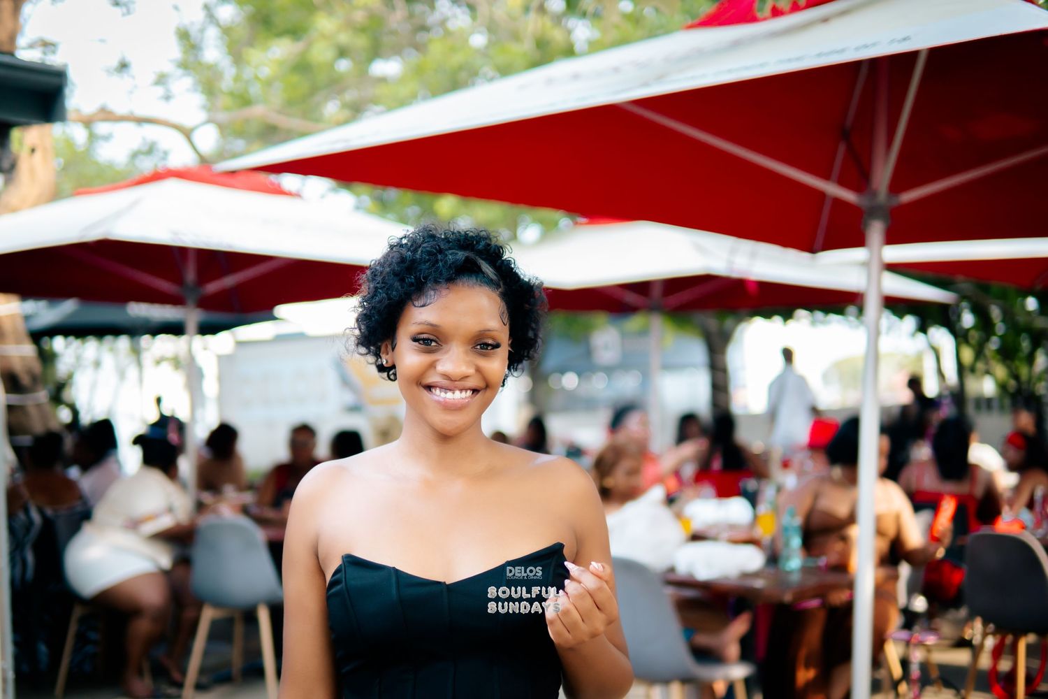 Smiling woman in black top holds a card at an outdoor restaurant with red umbrellas and guests in background.