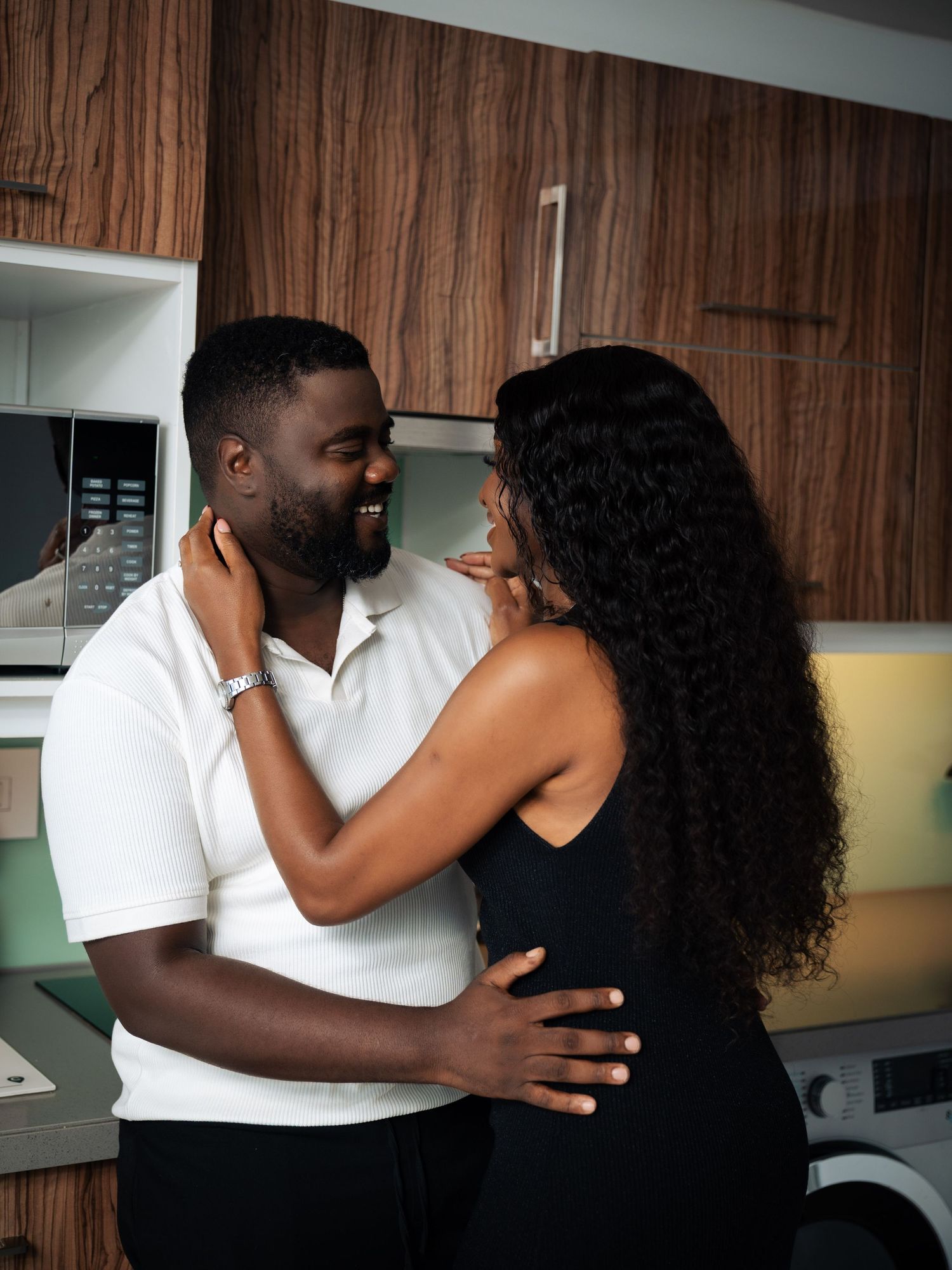 A couple embraces and smiles at each other in a modern kitchen, woman in black dress, man in white polo shirt.