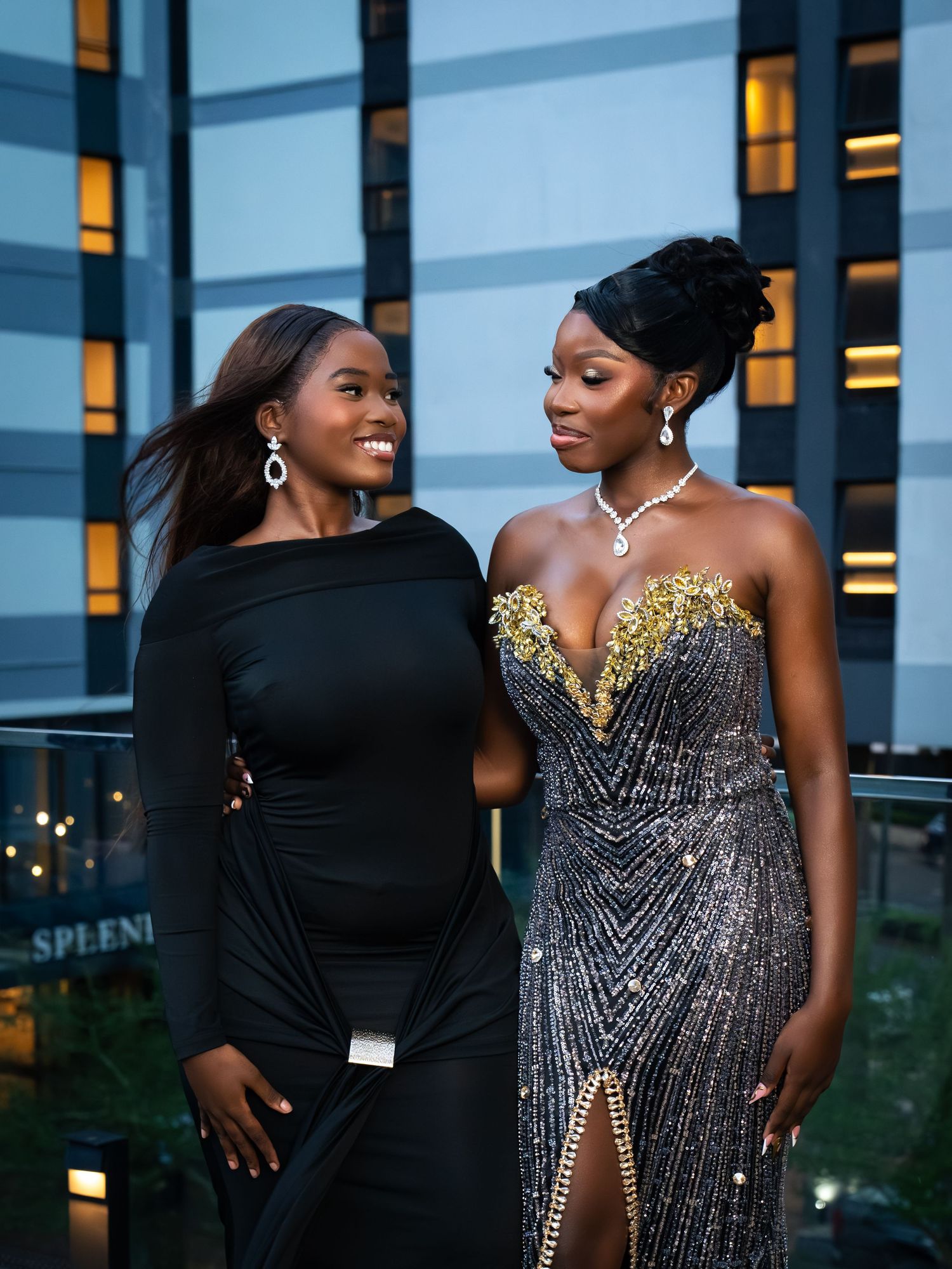 Two elegant women in black formal gowns pose outdoors at dusk near a modern hotel building.