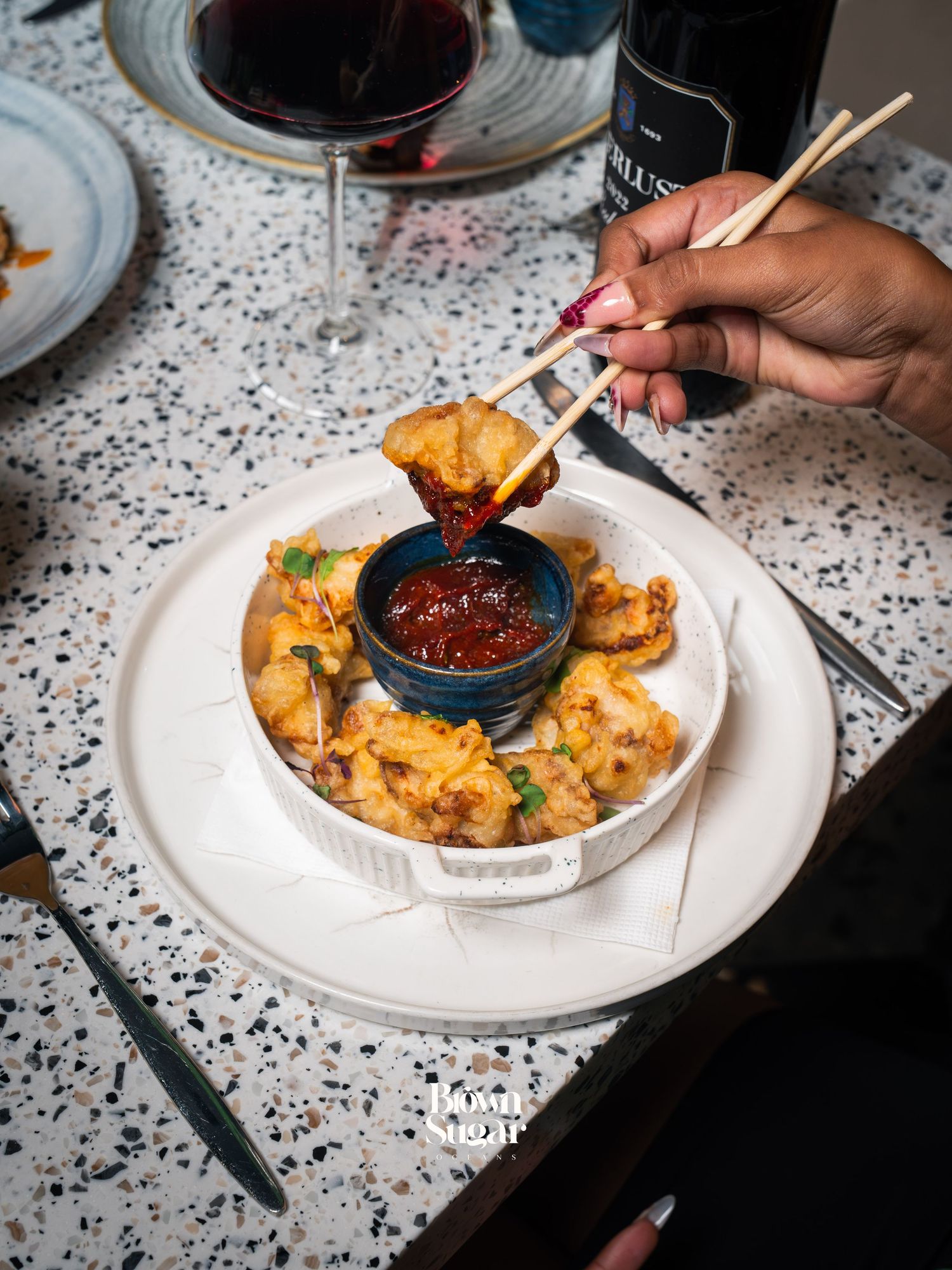 Hand using chopsticks to dip crispy fried dumpling into spicy sauce, served with red wine on terrazzo table.