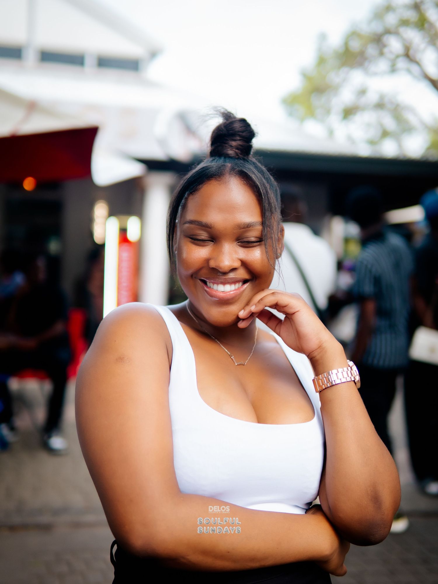 A smiling young Black woman in a white crop top with her hair up, posing outdoors at a casual event.
