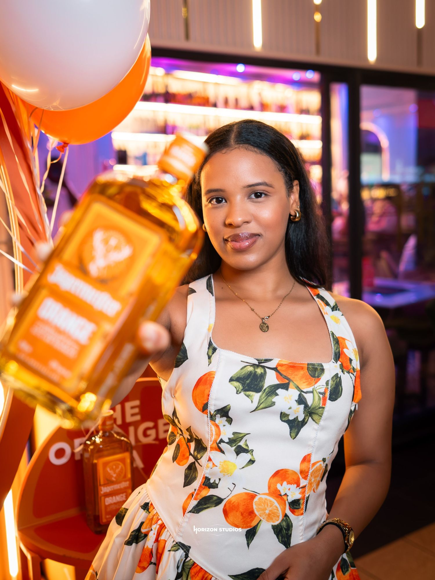 Woman in floral dress holding a bottle of spirits at a vibrant orange-themed event with balloons.