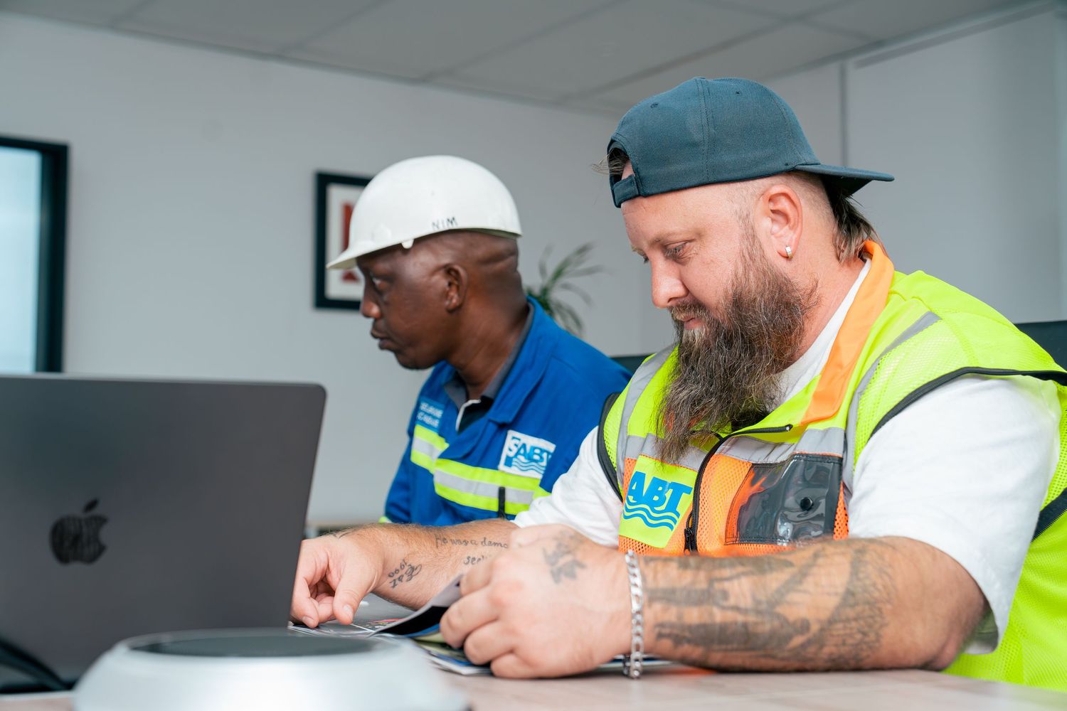 Two construction workers in safety vests review project details on a laptop computer at a desk indoors.