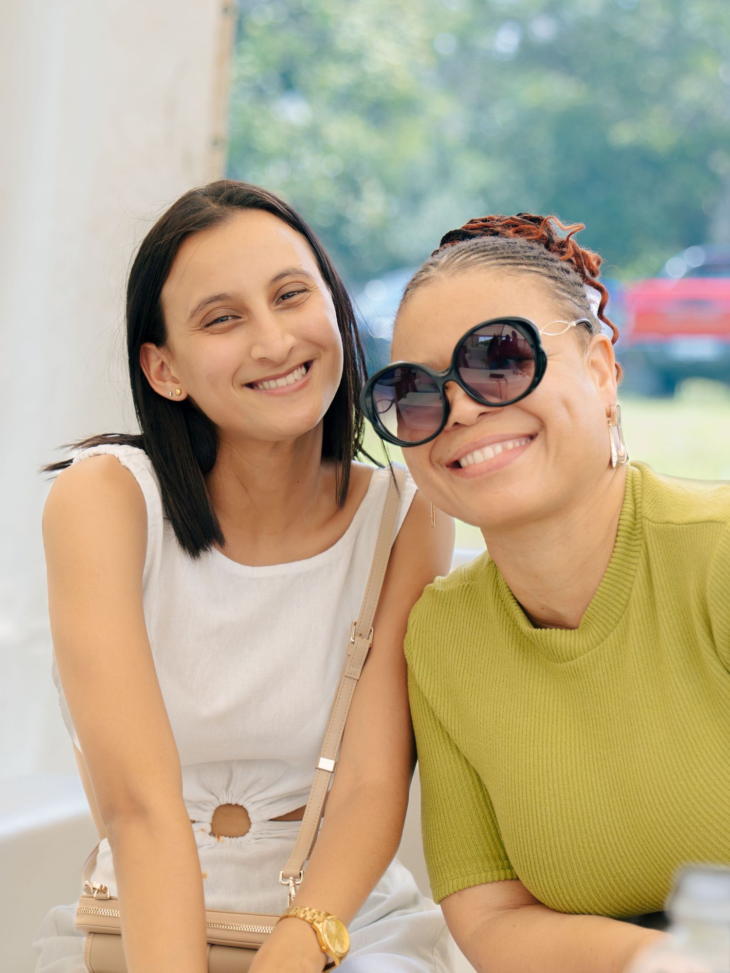 Two smiling women posing together outdoors, one in white outfit and one in green top wearing large sunglasses.